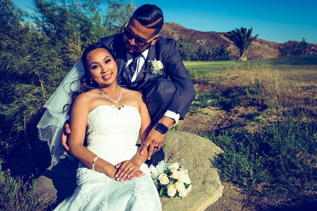 A newlywed couple outdoors with mountains and trees in the background, the bride is sitting in a white wedding dress with a veil and jewelry, and the groom is kneeling beside her in a dark suit with sunglasses, both smiling.