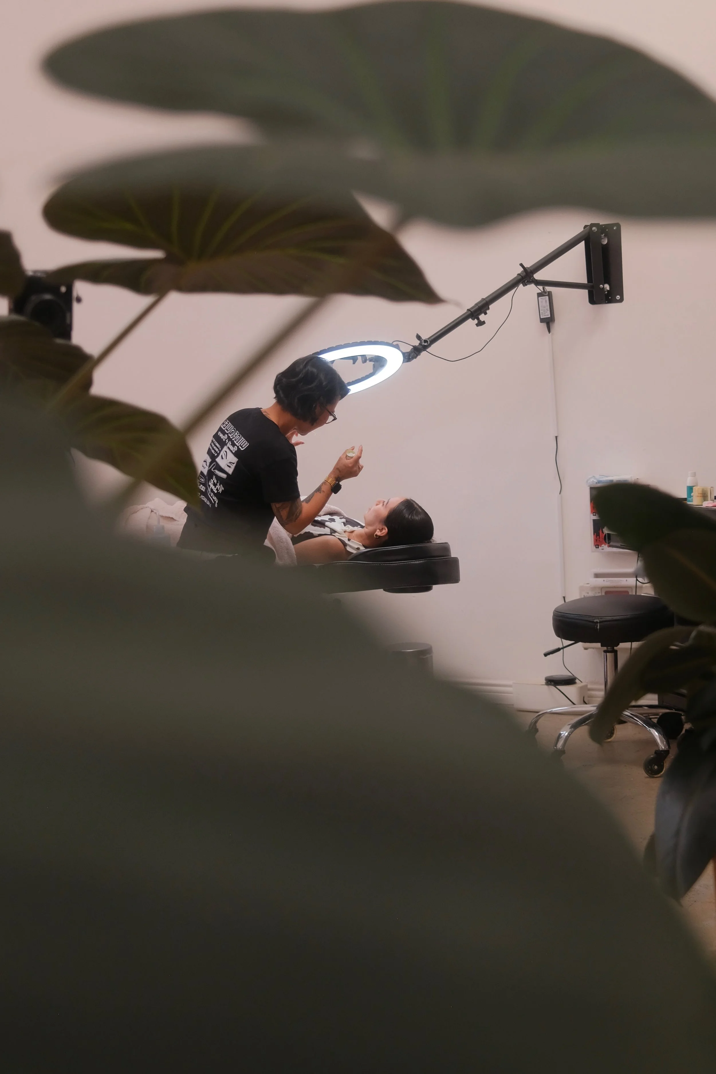 A person lying down receiving a procedure from Rachelle, with large green leaves partially obscuring the view.