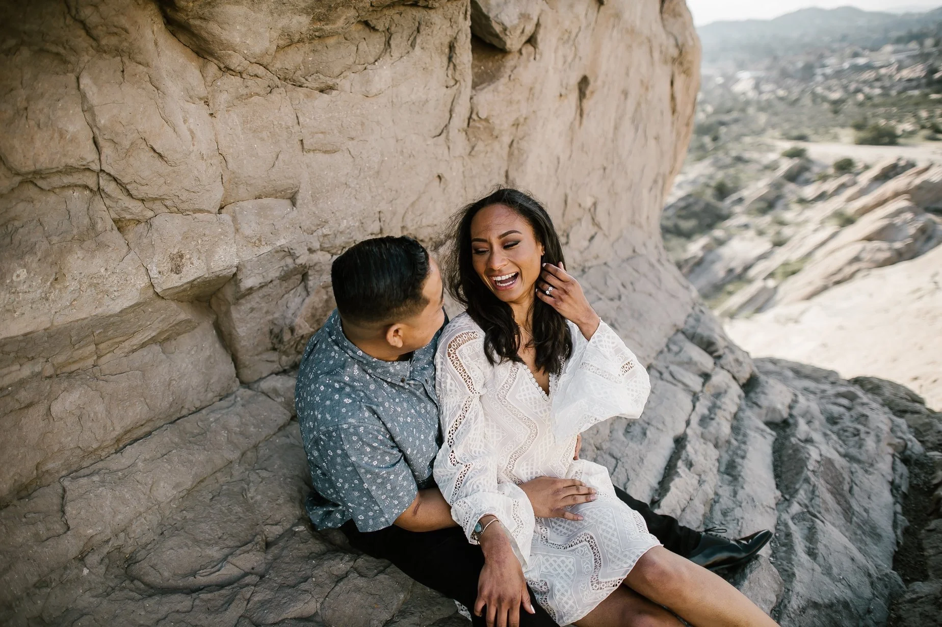 A man and woman sitting on rocks in a desert landscape, smiling and looking at each other. The woman is wearing a white dress, and the man is wearing a blue patterned shirt.