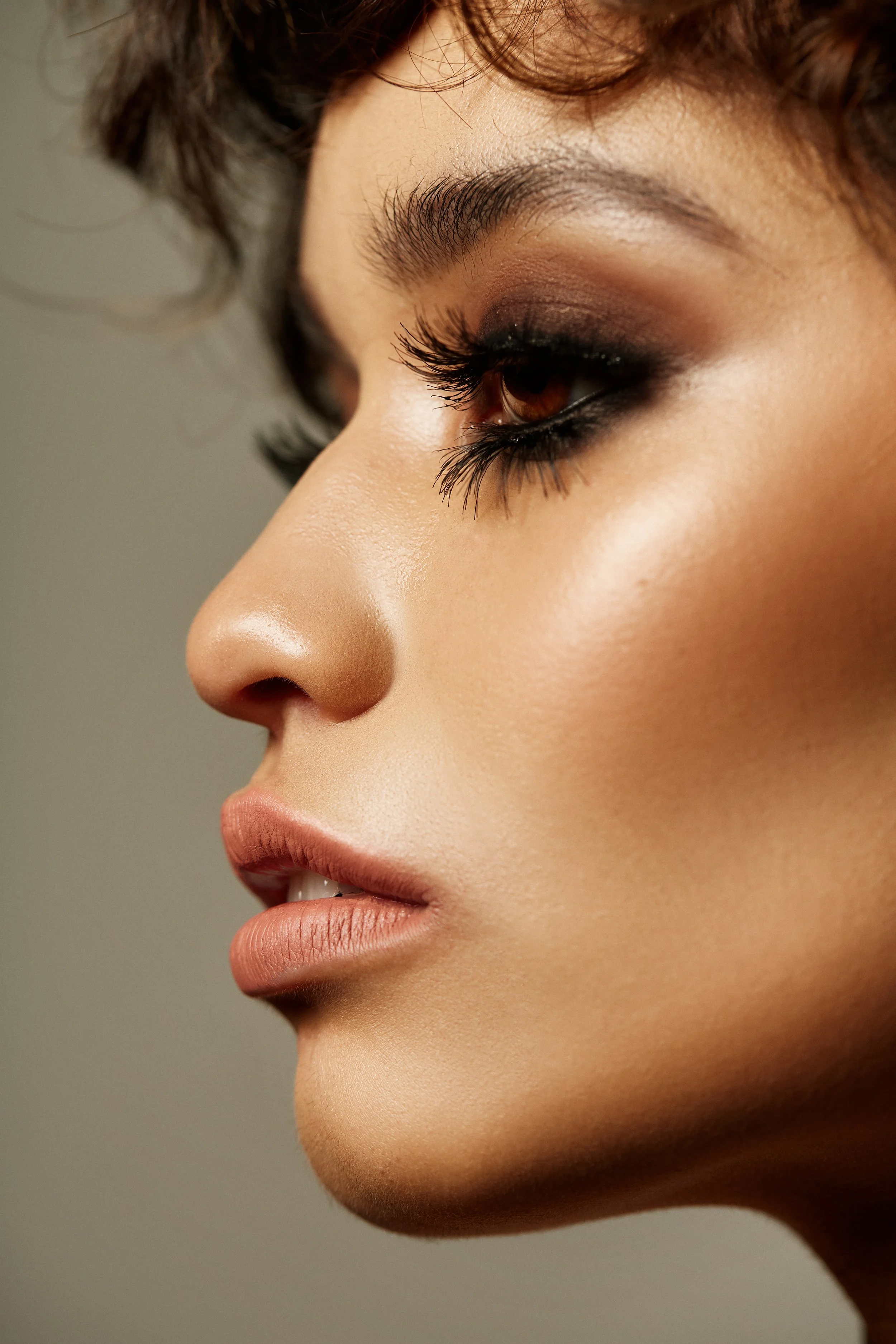 Close-up side view of a woman's face showcasing makeup with smoky eye shadow, long eyelashes, smooth skin, and natural lip color.