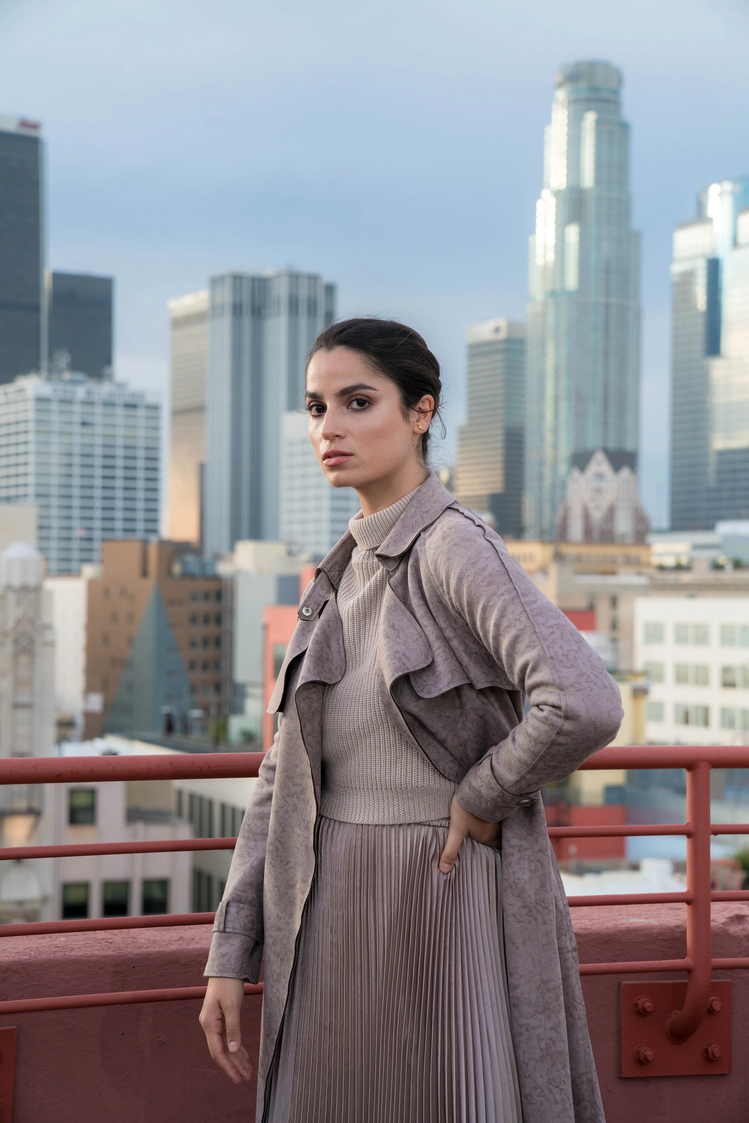 A young woman with dark hair styled back, wearing a light beige turtleneck, taupe jacket, and pleated skirt, stands on a city rooftop with a cityscape of tall buildings in the background.