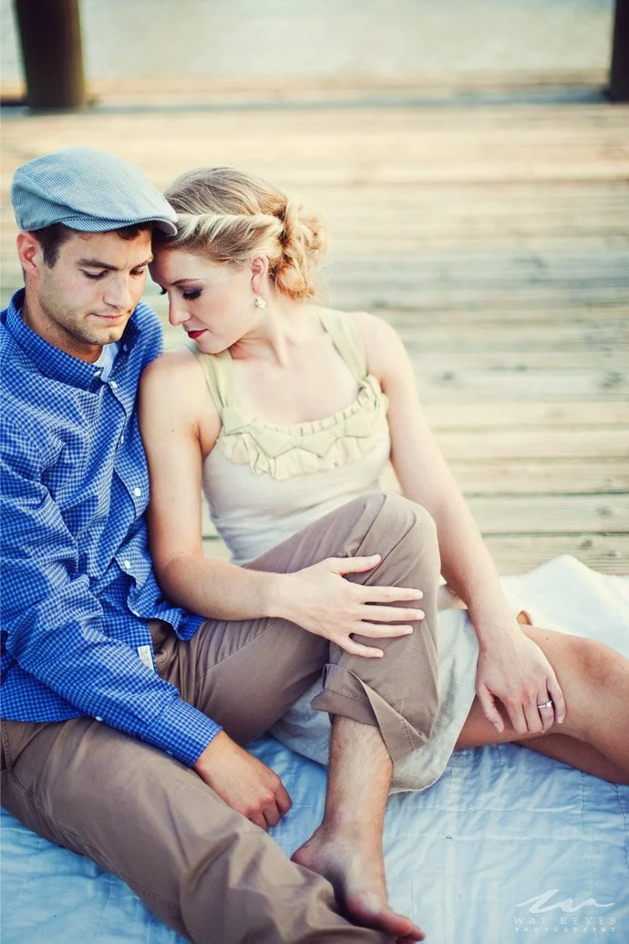 A man and woman sitting close together on a blanket on a wooden deck, with their heads touching in an intimate pose.