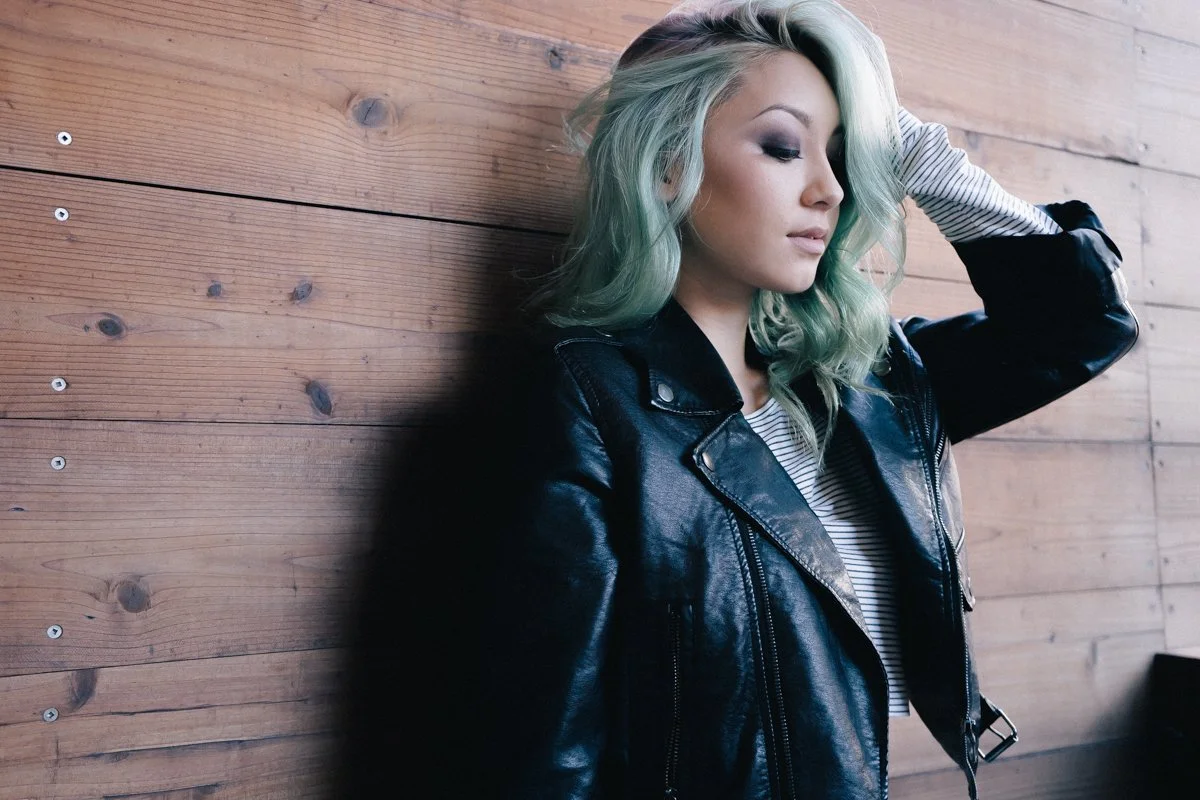 A young woman with pastel green hair leaning against a wooden wall, wearing a black leather jacket and a striped shirt, with her eyes closed and her hand in her hair.