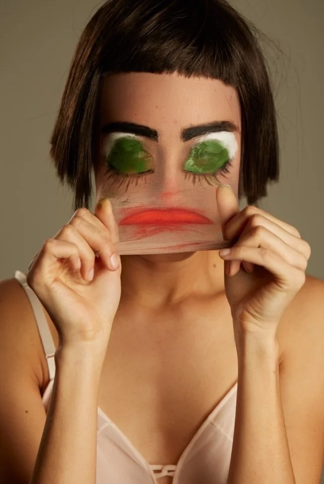 Person holding a transparent mask with a painted face, including green eyes, long eyelashes, thick black eyebrows, and red lips, in front of their face.