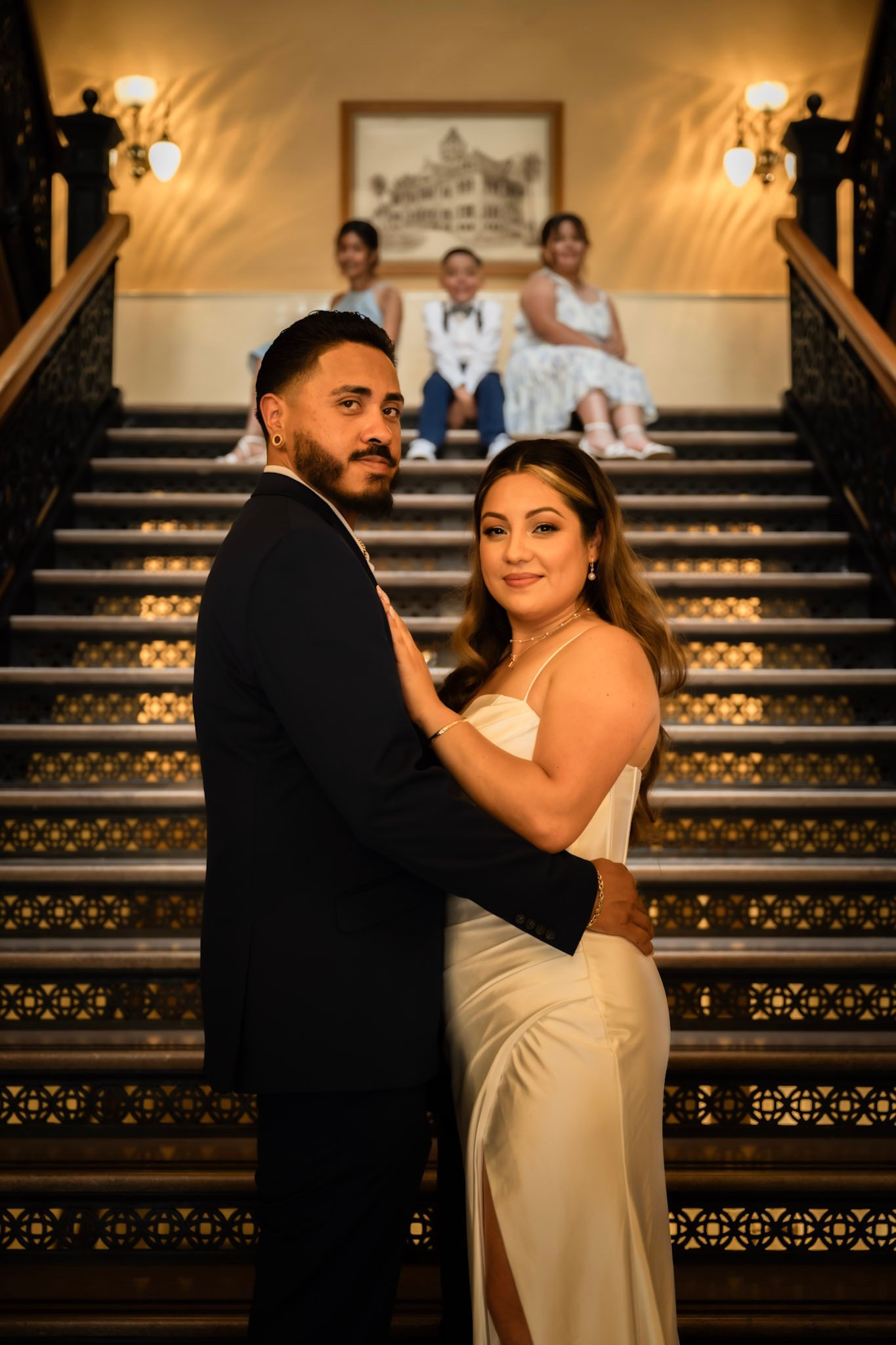 A couple dressed in formal attire standing on ornate stairs in front of a warm-toned wall with wall sconces and a framed picture. People are sitting on the stairs behind them.