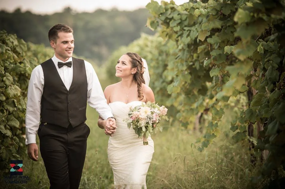 A newlywed couple walking hand-in-hand through a vineyard, with the groom dressed in a black vest and bow tie, and the bride holding a bouquet of flowers, smiling at each other.