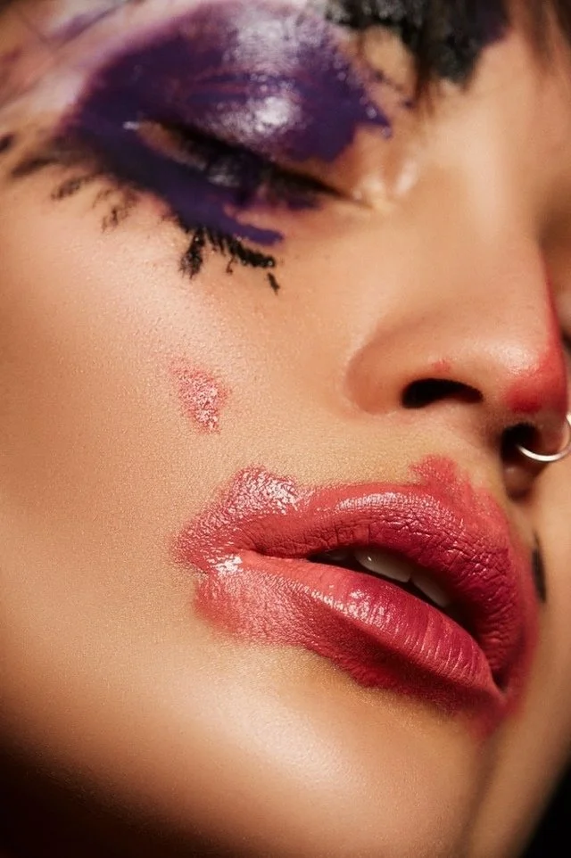 Close-up of a person's face showing purple eye makeup, red lipstick, and a small facial blemish.