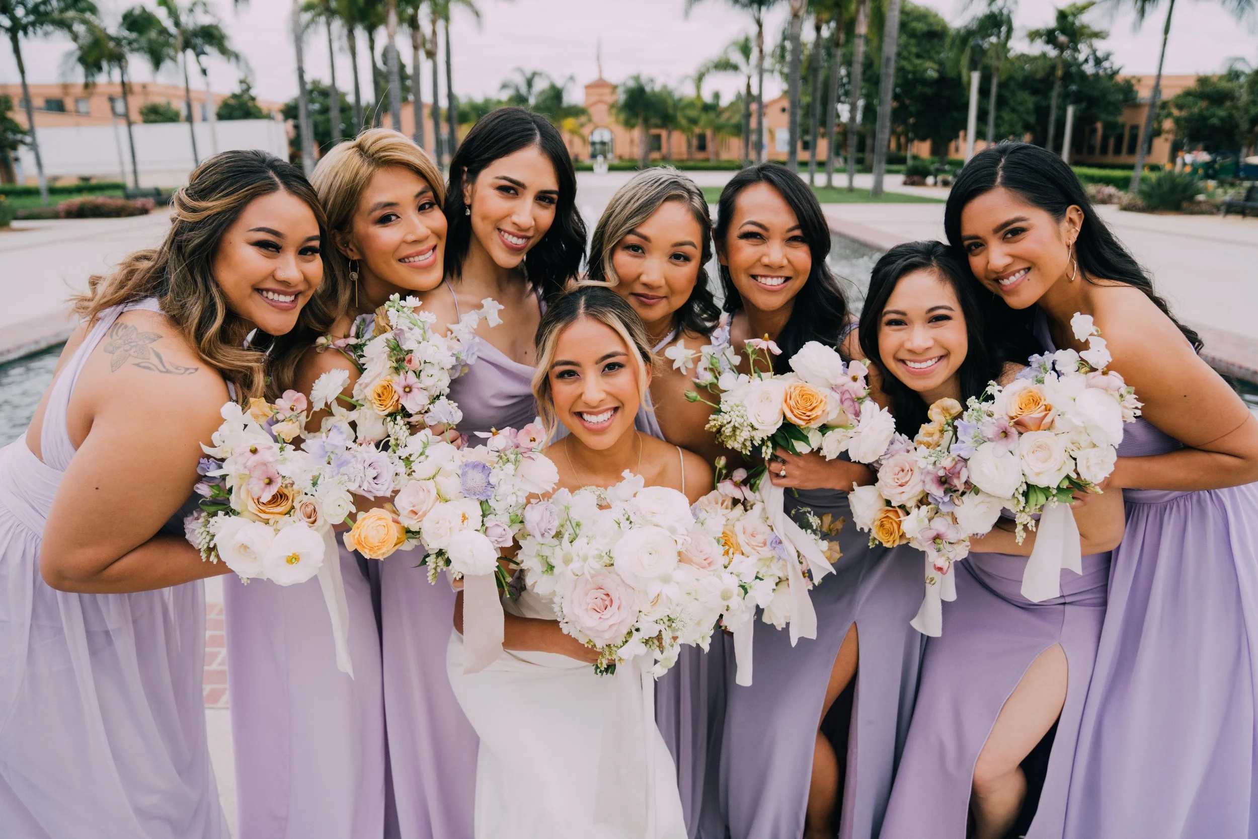 Group of women dressed in lavender bridesmaid dresses holding bouquets of flowers, smiling outdoors.
