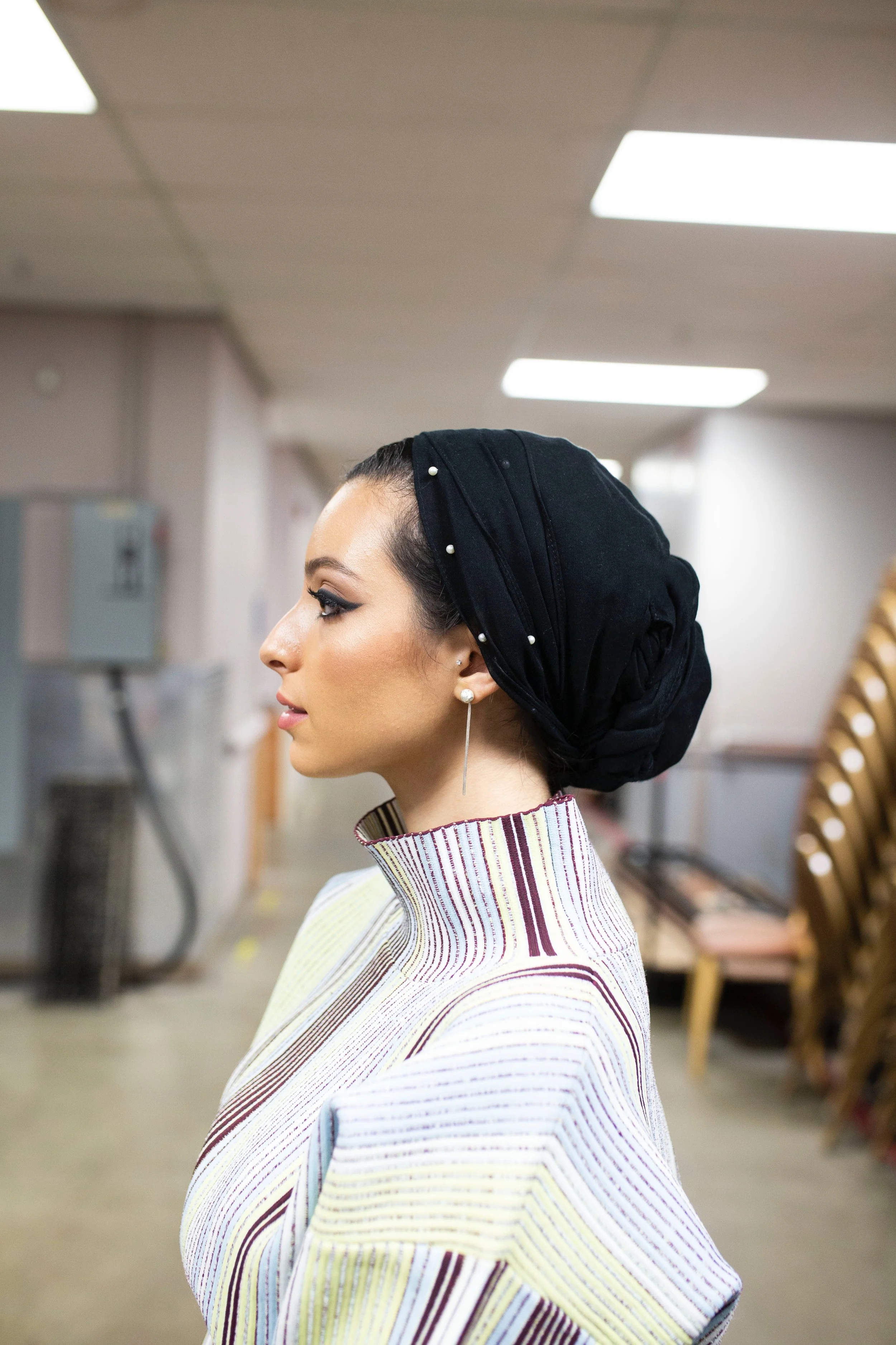 A woman with dark hair styled in a bun wearing a black headscarf with pearl embellishments, earrings, and a colorful striped dress, standing in an indoor setting with chairs and a hallway.