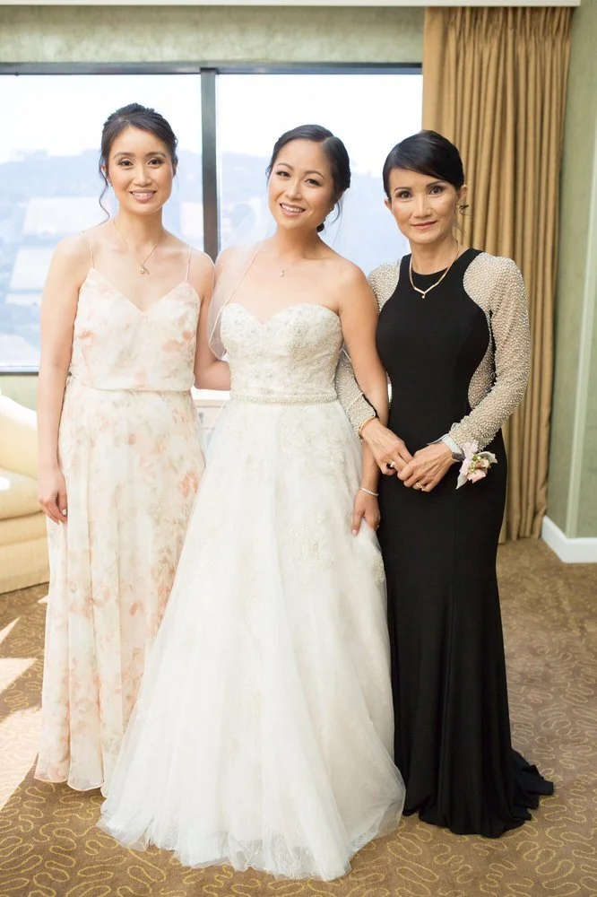Three women standing indoors in front of a window with yellow curtains, one in a wedding gown and the other two in formal dresses, smiling at the camera.