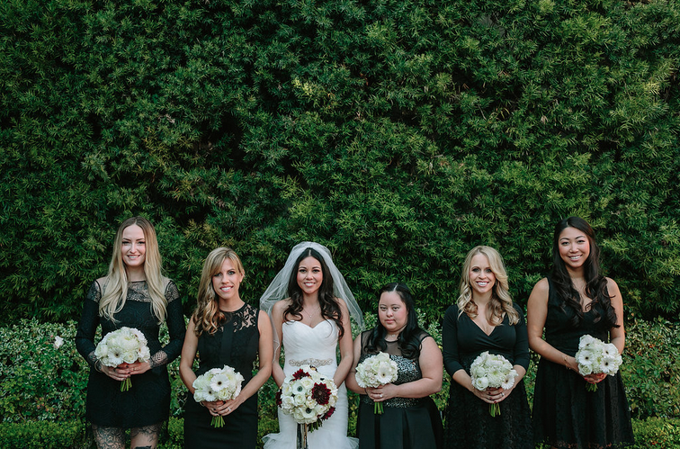 Group of six women, including a bride in a white wedding dress and veil, holding bouquets of white and burgundy flowers, standing against a lush green hedge background.