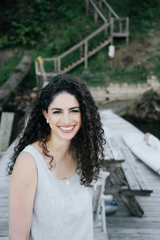 A young woman with long curly hair smiling outdoors near a wooden dock with trees and stairs in the background.