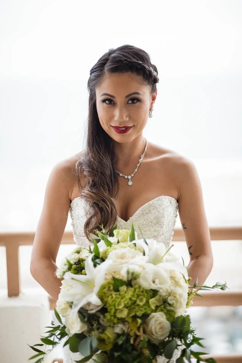 A bride in a strapless wedding dress holding a bouquet of white and green flowers, smiling at the camera.