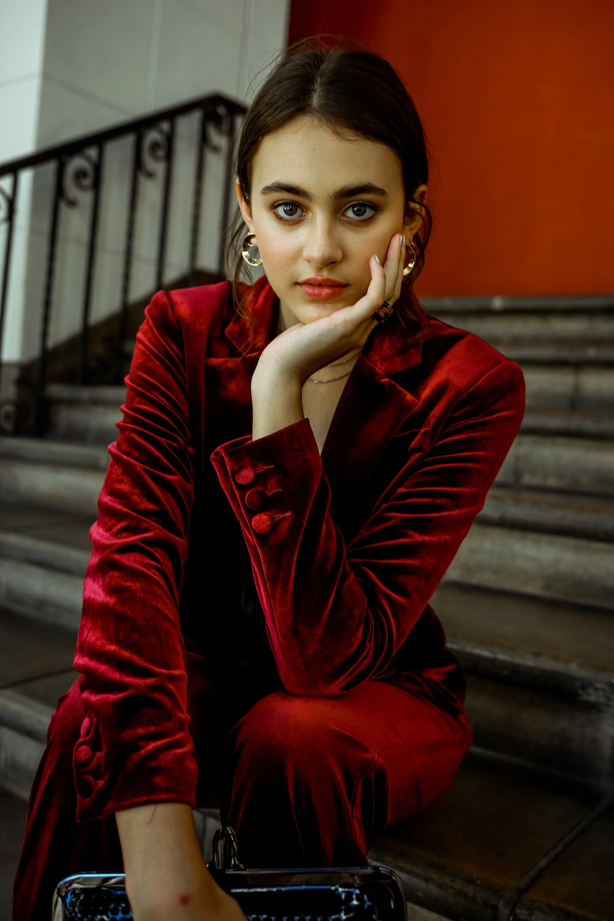 A young woman with blue eyes and dark brown hair, wearing a red velvet blazer, sitting on stairs with her hand resting on her cheek, looking directly at the camera. The background features a reddish wall and black metal railing.