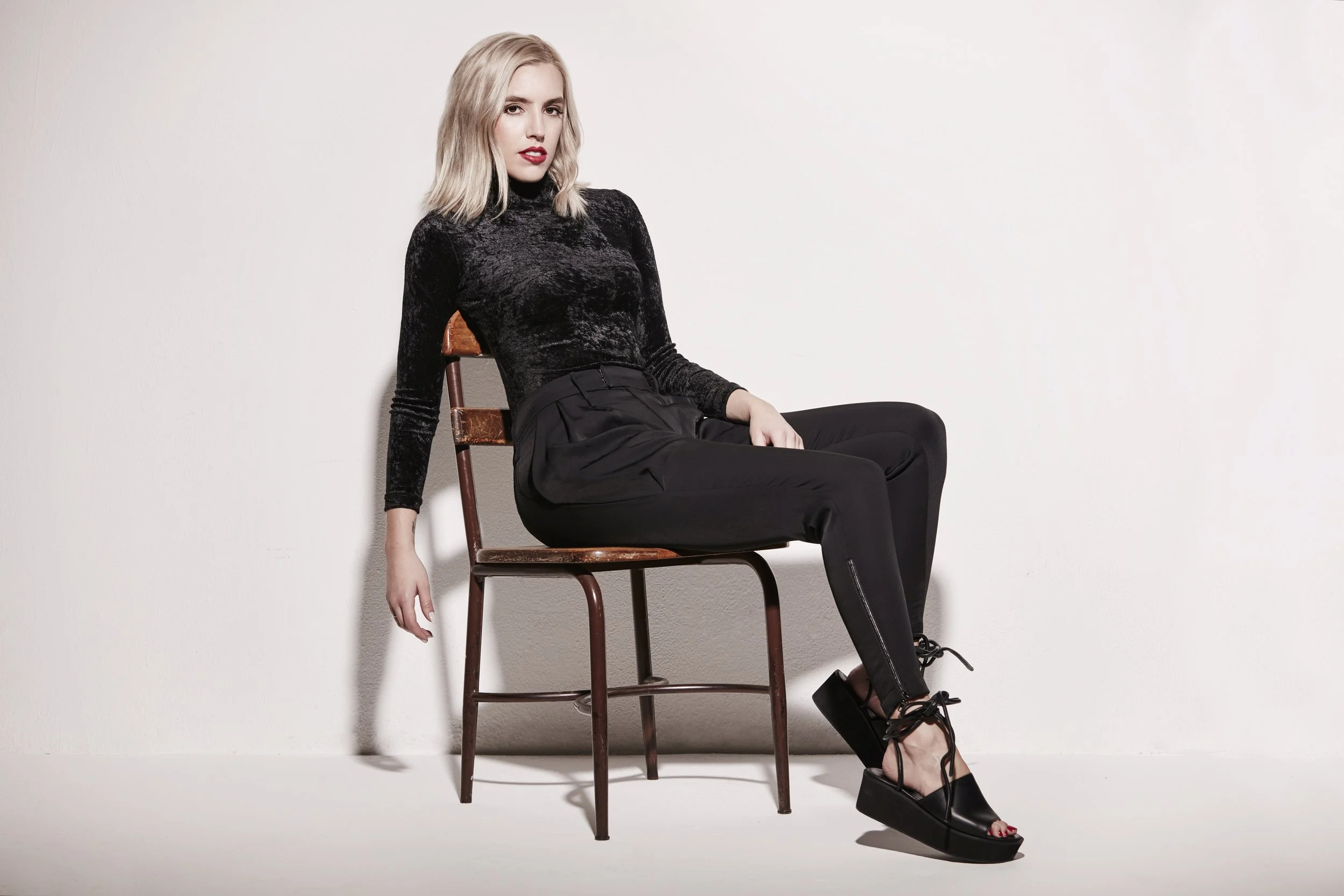 A young woman with blonde hair sitting sideways on a wooden chair, wearing a black velvet long-sleeve top, black high-waisted pants, and black platform shoes with laces, against a plain white background.
