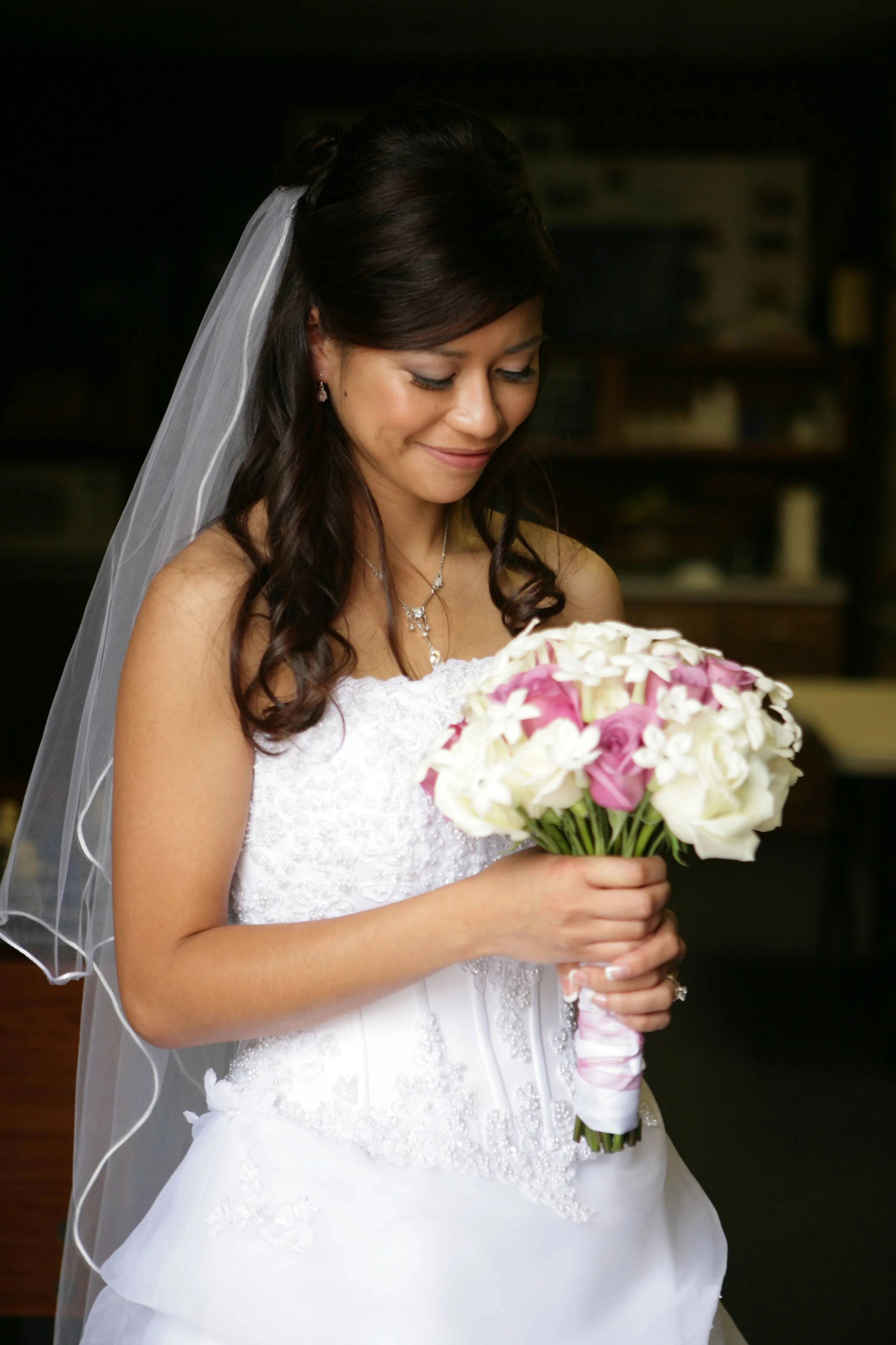 A smiling bride with dark brown hair in a wedding dress, holding a bouquet of pink and white roses, wearing earrings, a necklace, and a veil.