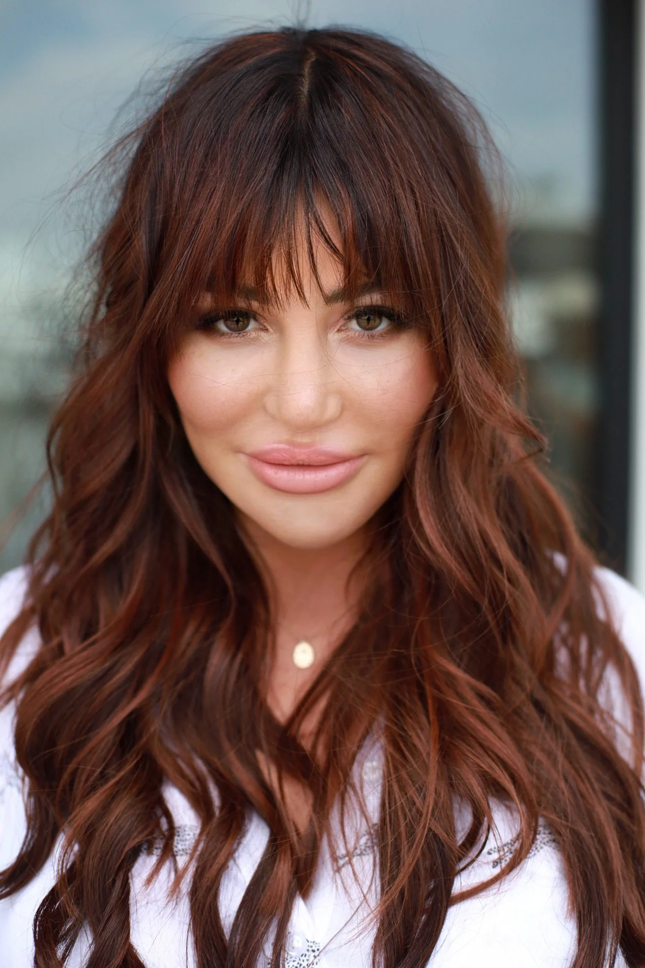 Close-up of a woman with long, wavy auburn hair and bangs, smiling softly, wearing a white top and a necklace.