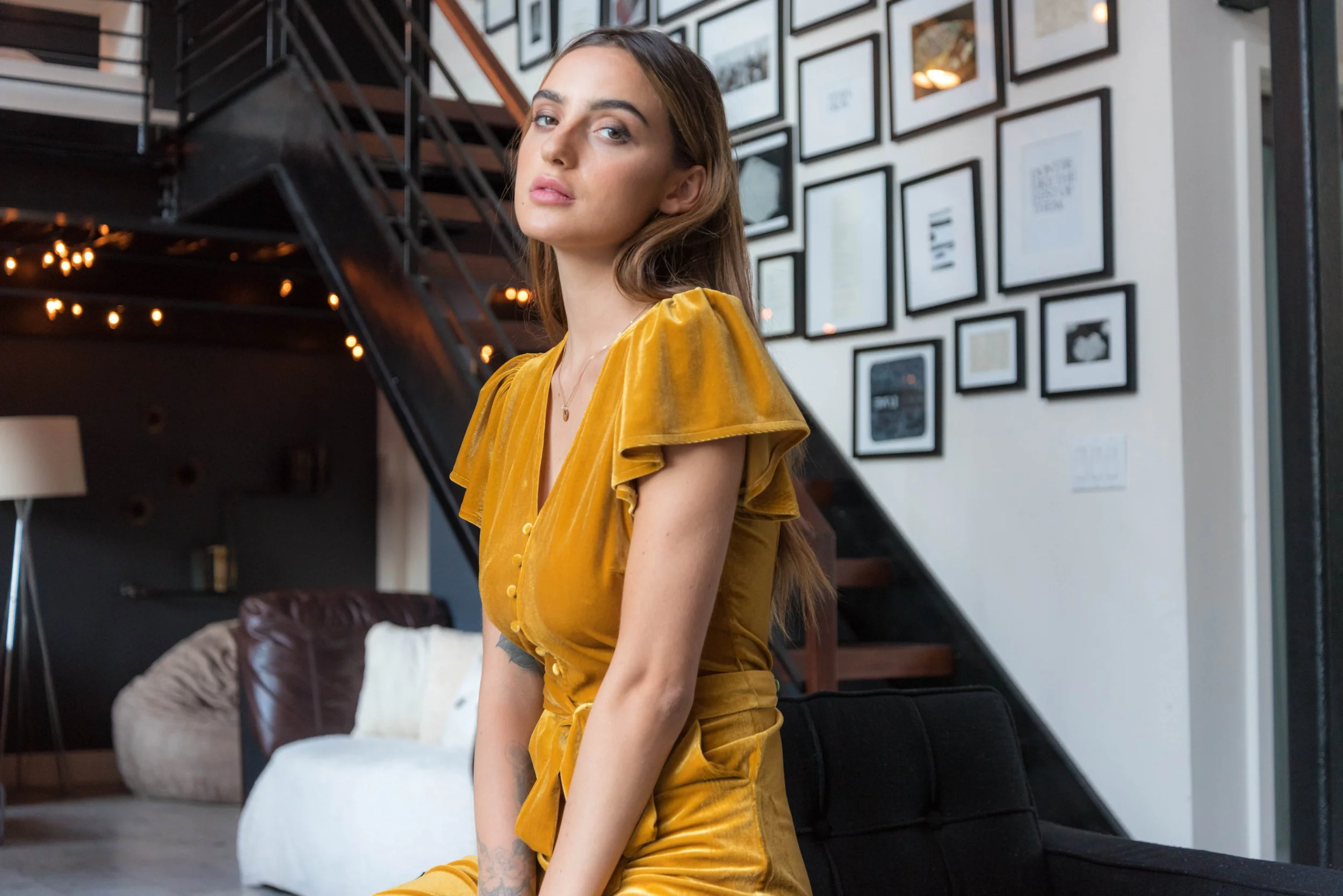A young woman with long brown hair and light skin, wearing a yellow satin blouse and matching skirt, sitting on a black chair in a modern interior with framed photos on the wall.