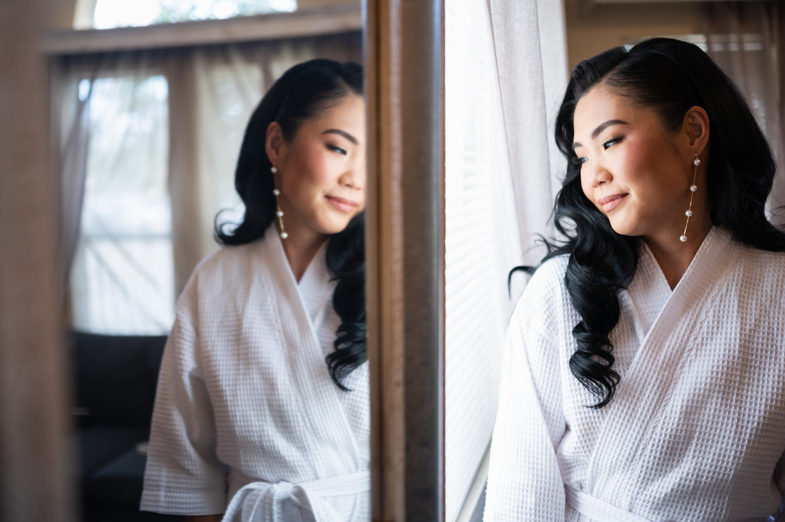 A woman with long black wavy hair wearing pearl earrings and a light-colored robe looks at her reflection in a mirror, with soft sunlight coming through a window beside her.