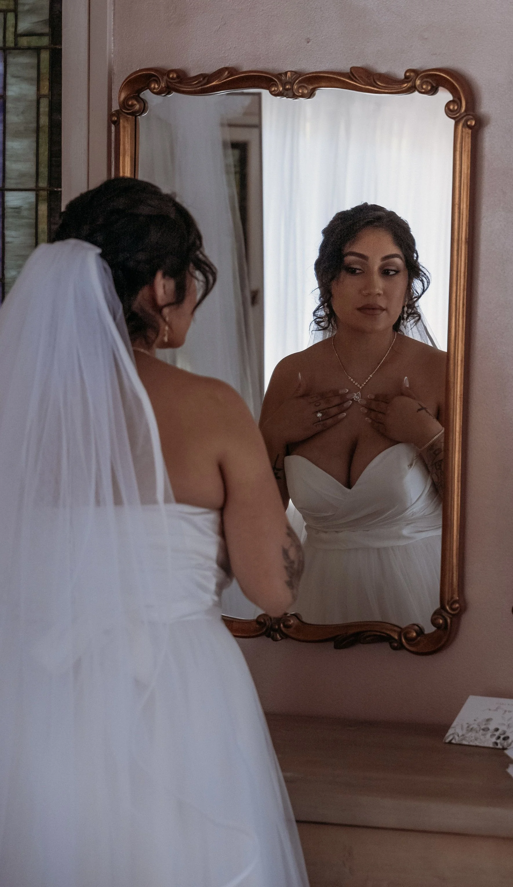 A woman in a white wedding dress looks at herself in an ornate wooden framed mirror, adjusting her necklace, with her reflection showing her face and upper body, in a softly lit room.