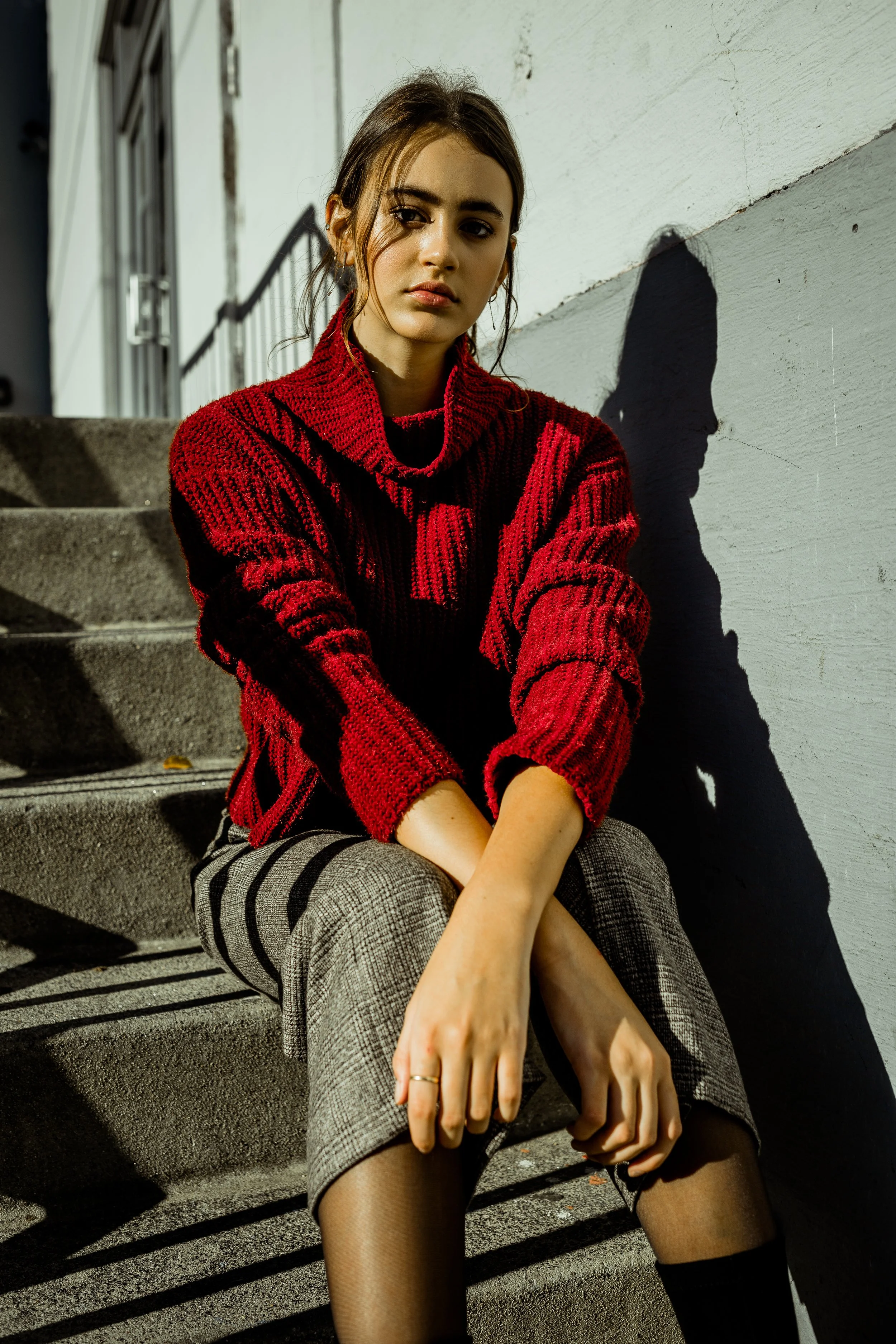 A young woman with brown hair in a loose updo and light makeup sits on outdoor concrete stairs, wearing a red and black striped turtleneck sweater, plaid skirt, and black sheer stockings, with sunlight casting a shadow of her profile on the white wal