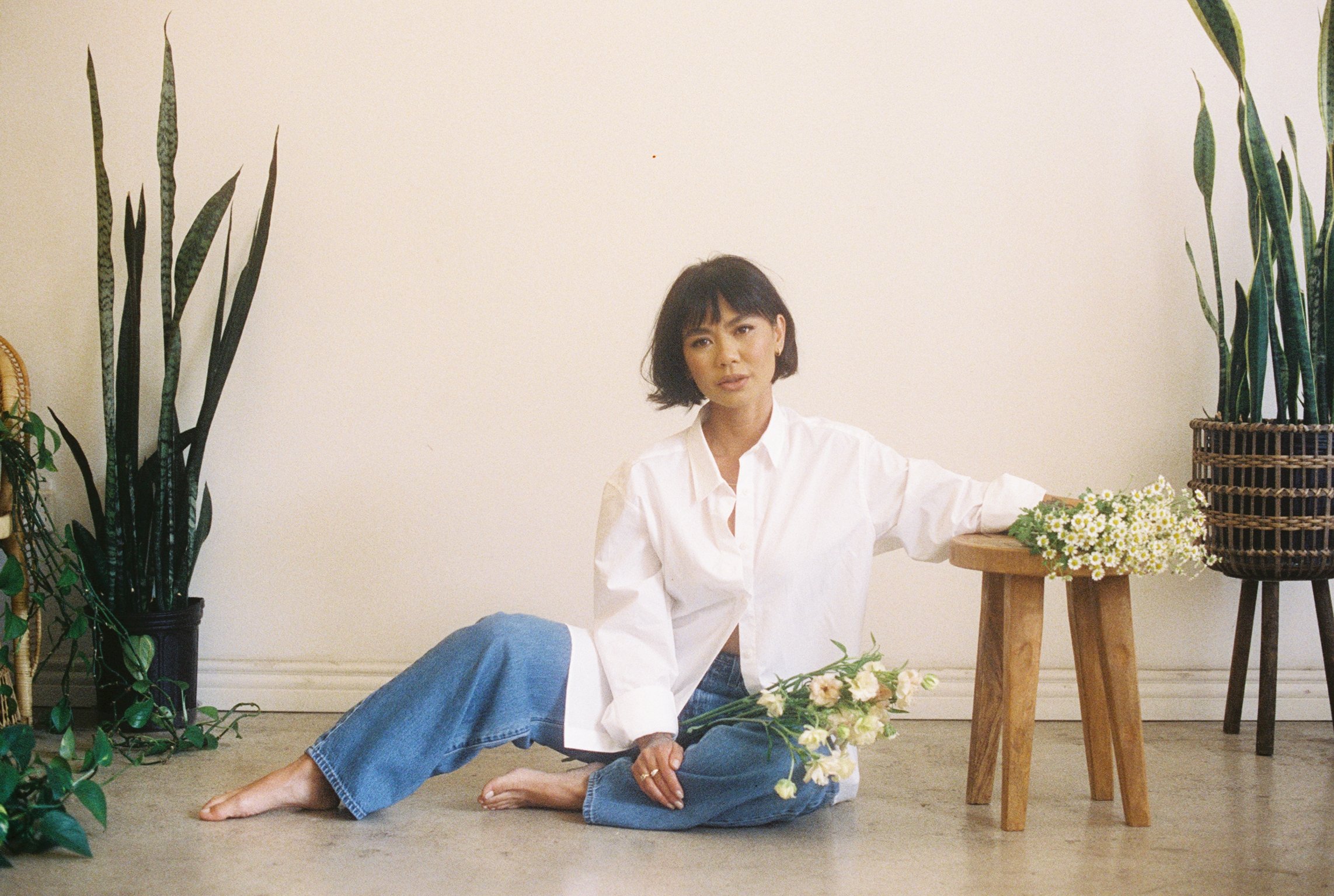 Rachelle short dark hair, wearing a white shirt and blue jeans, sitting on the floor next to a bouquet of daisies. She is indoors, surrounded by potted plants and wooden furniture, against a plain off-white wall.