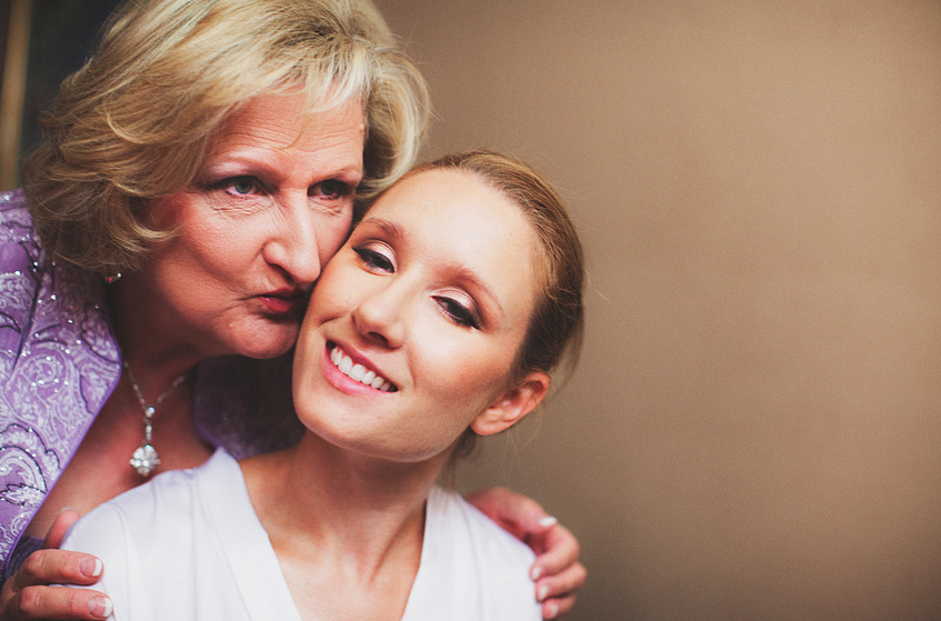 An older woman wearing a purple dress is giving a kiss on the cheek to a young woman with light brown hair, who is smiling.