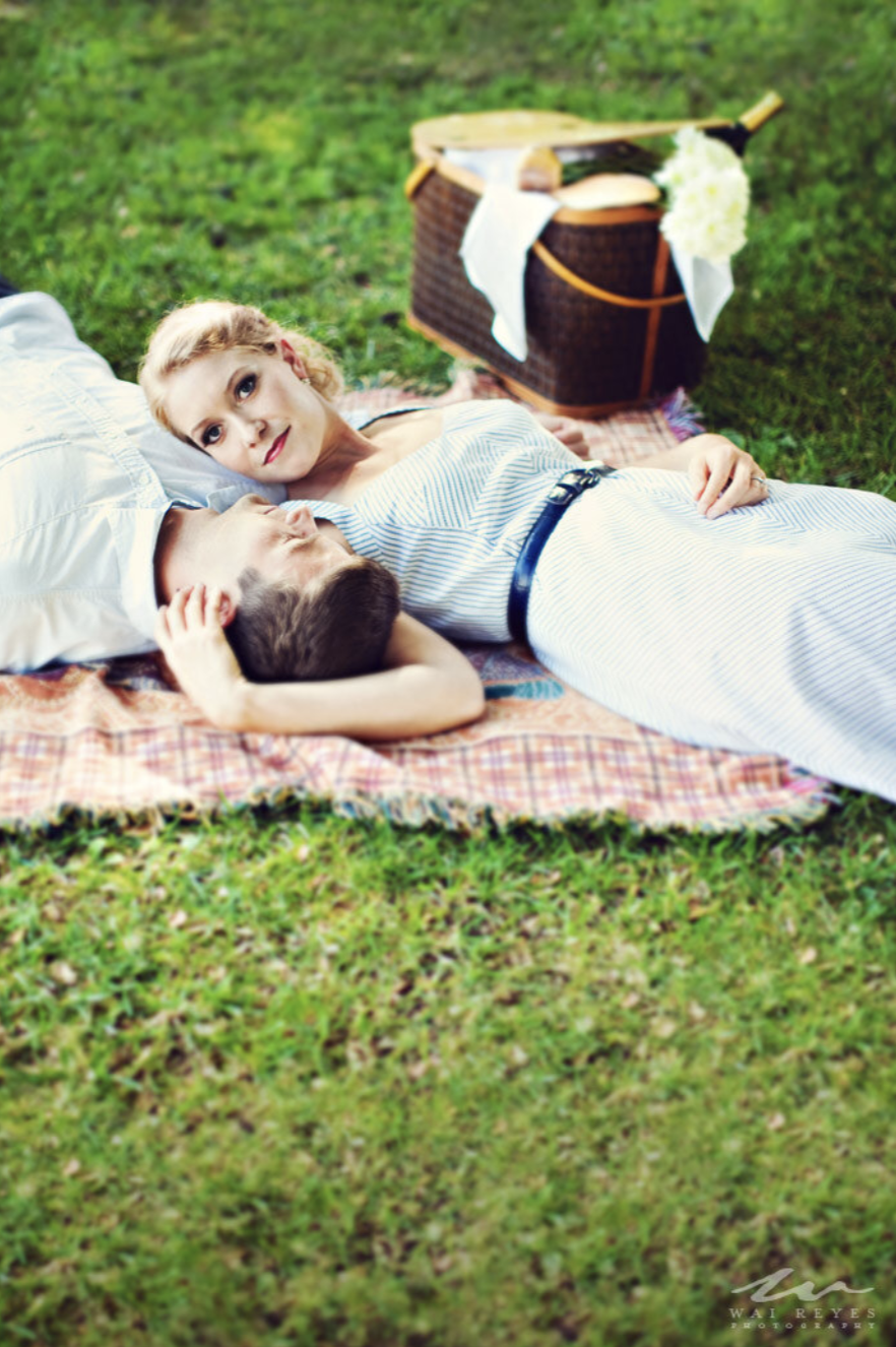 A woman and a young girl are lying on a blanket on the grass, with the woman resting her head on a pillow and the girl looking at her, beside a picnic basket.
