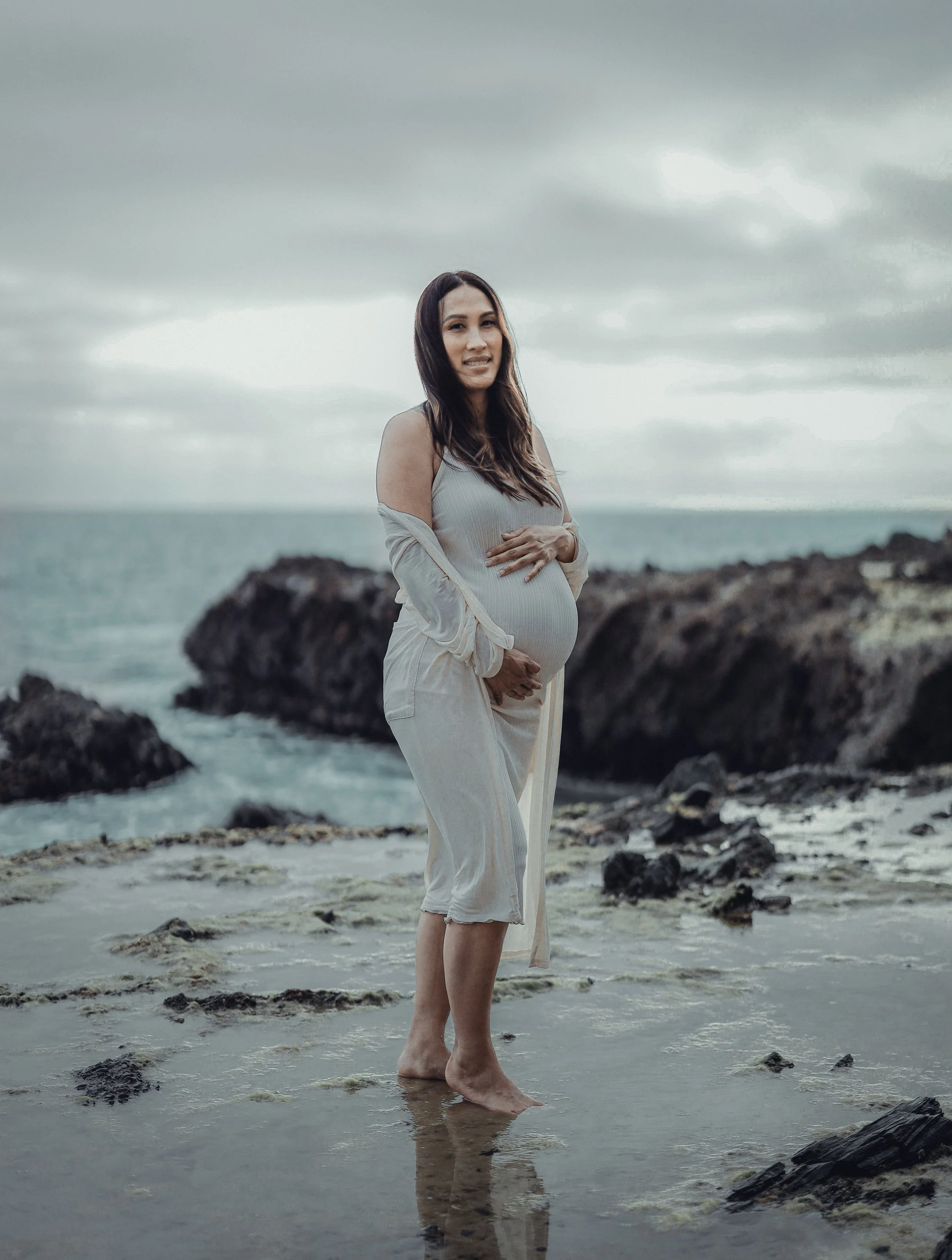 A pregnant woman standing barefoot on the beach with rocky shoreline, holding her belly and smiling at the camera under a cloudy sky.