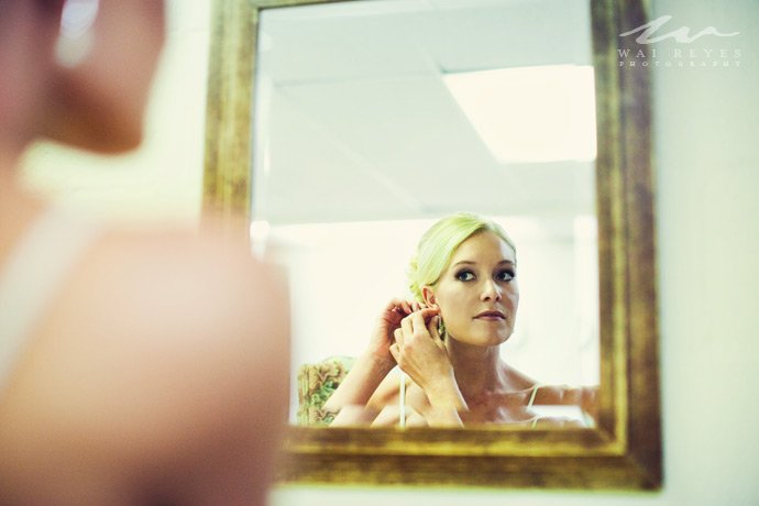 A woman with blonde hair, seen in a mirror, is adjusting her earring before a mirror.