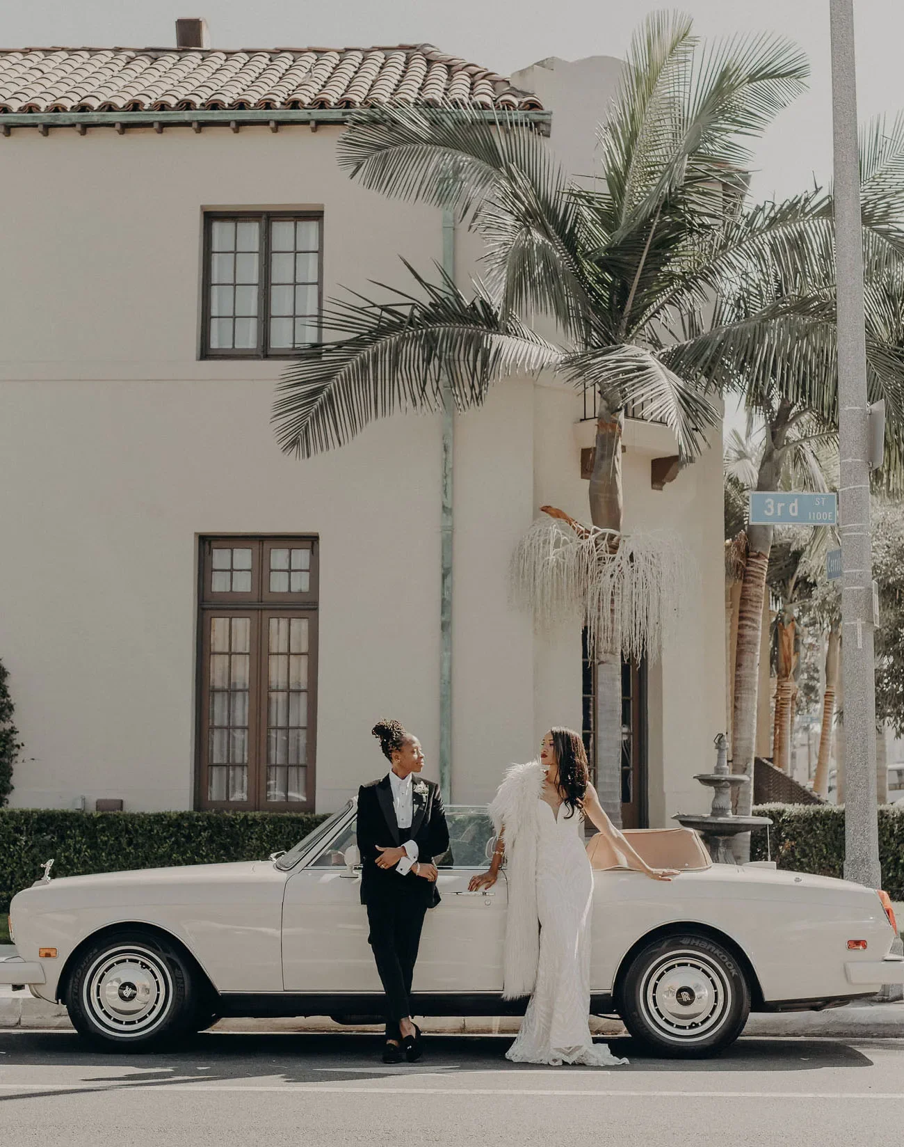 Two women in formal attire standing next to a white vintage convertible car parked on the street, with a beige building, palm trees, and street sign reading '3rd St' in the background.