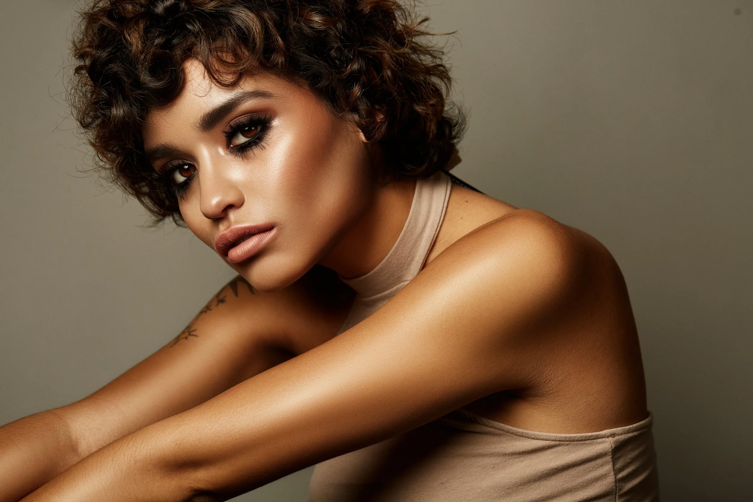 Close-up of a woman with curly brown hair and dark makeup, wearing a beige tank top, posing against a neutral background.