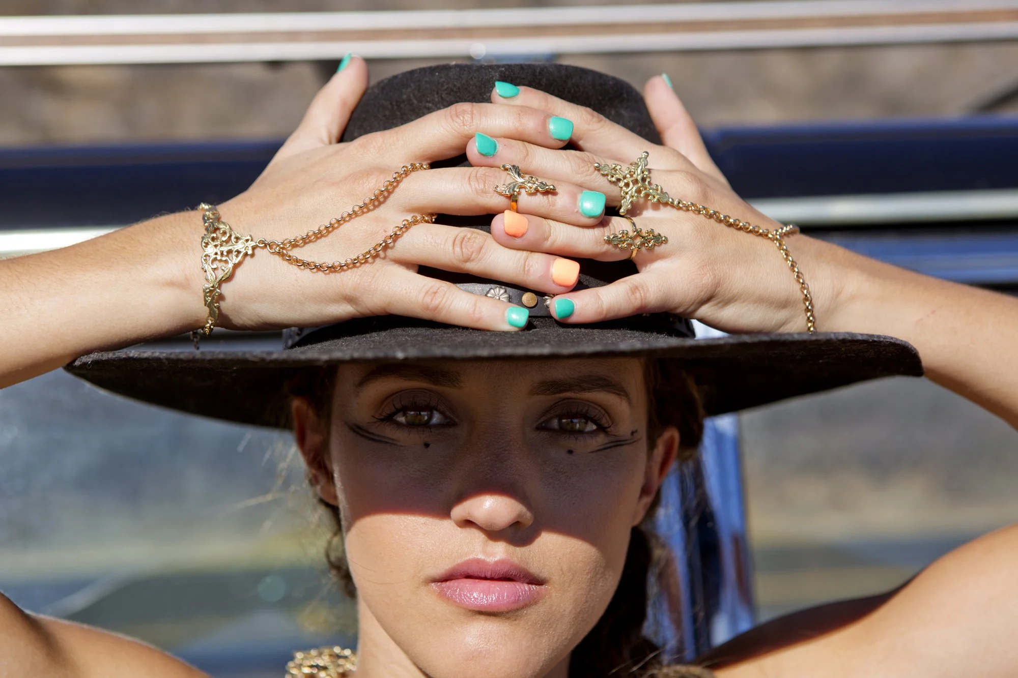 A woman wearing a wide-brimmed hat with jewelry, looking directly at the camera, outdoors with a sunny background.