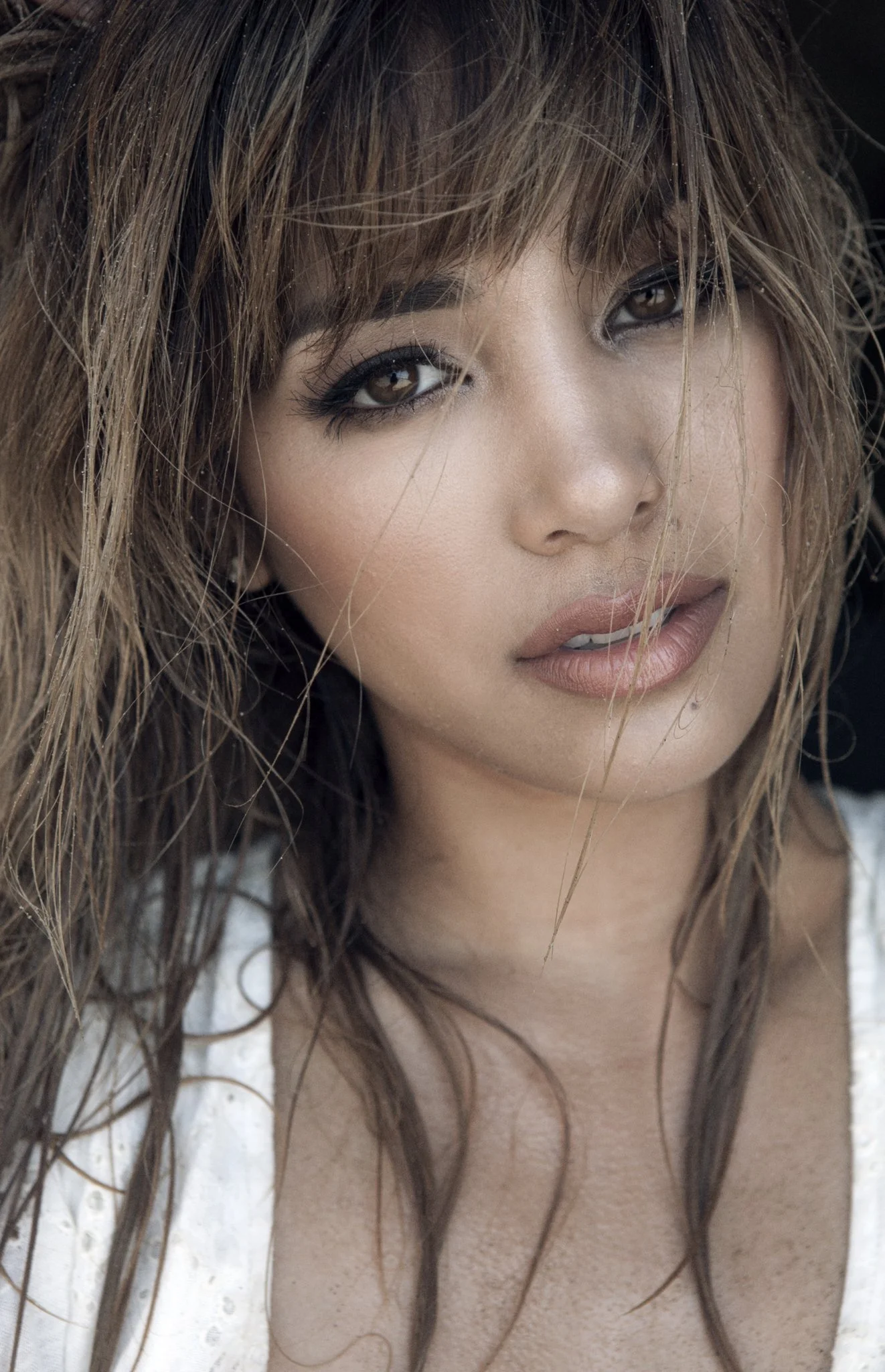 Close-up of a woman with light brown, wavy hair and brown eyes, wearing makeup, and a white garment with embroidery.