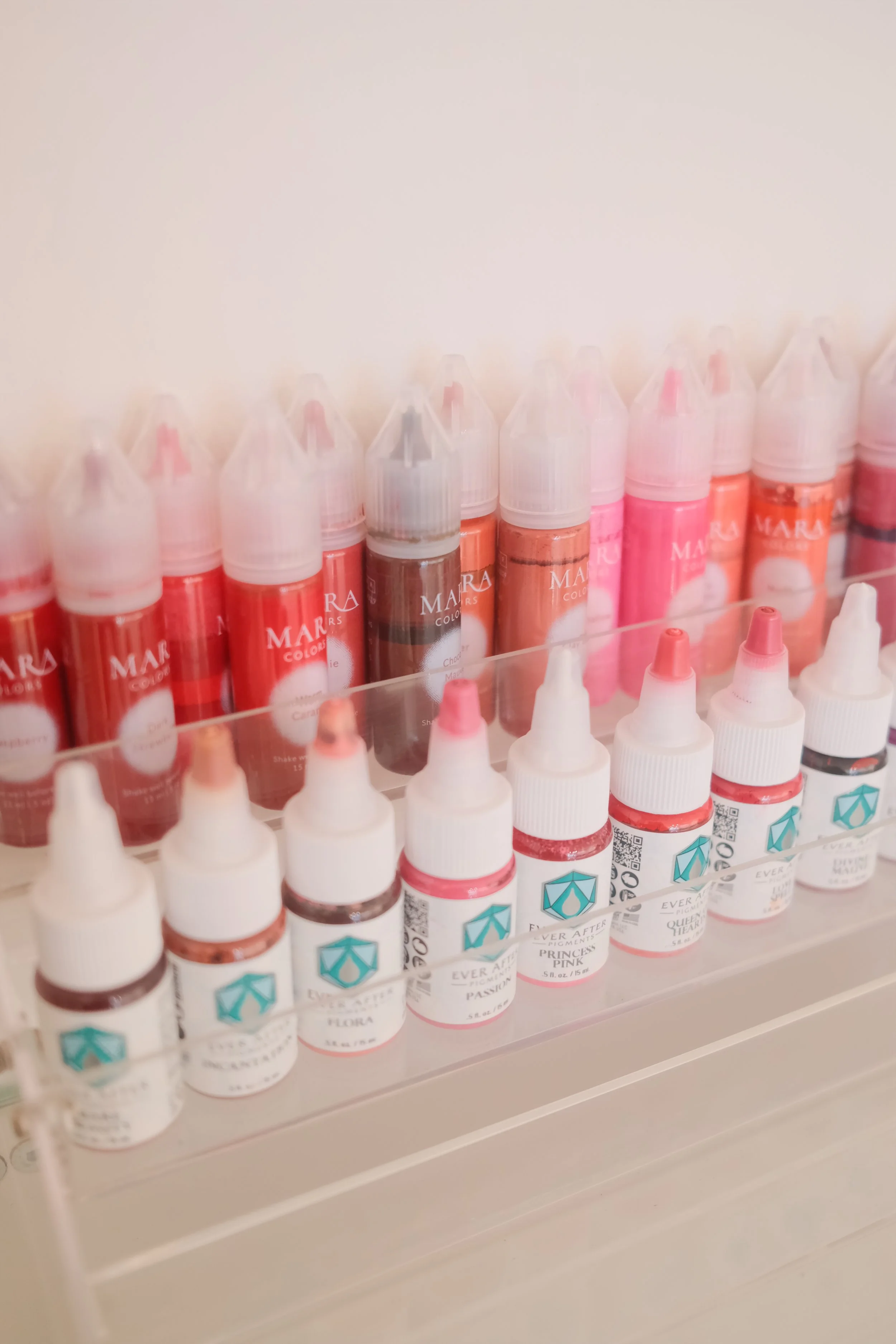 A variety of small bottles of liquid paint with different shades of pink, red, and brown, arranged on clear acrylic shelves.