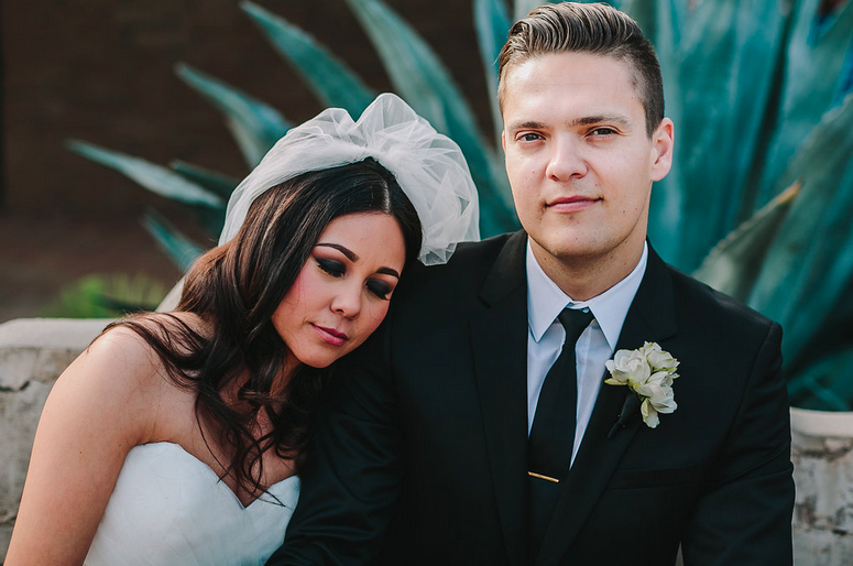 Bride and groom in wedding attire outdoors, with large green agave plants in the background. The bride has dark hair with loose waves, wearing a strapless white wedding dress and veil. The groom has short hair, wearing a black suit with a white shirt