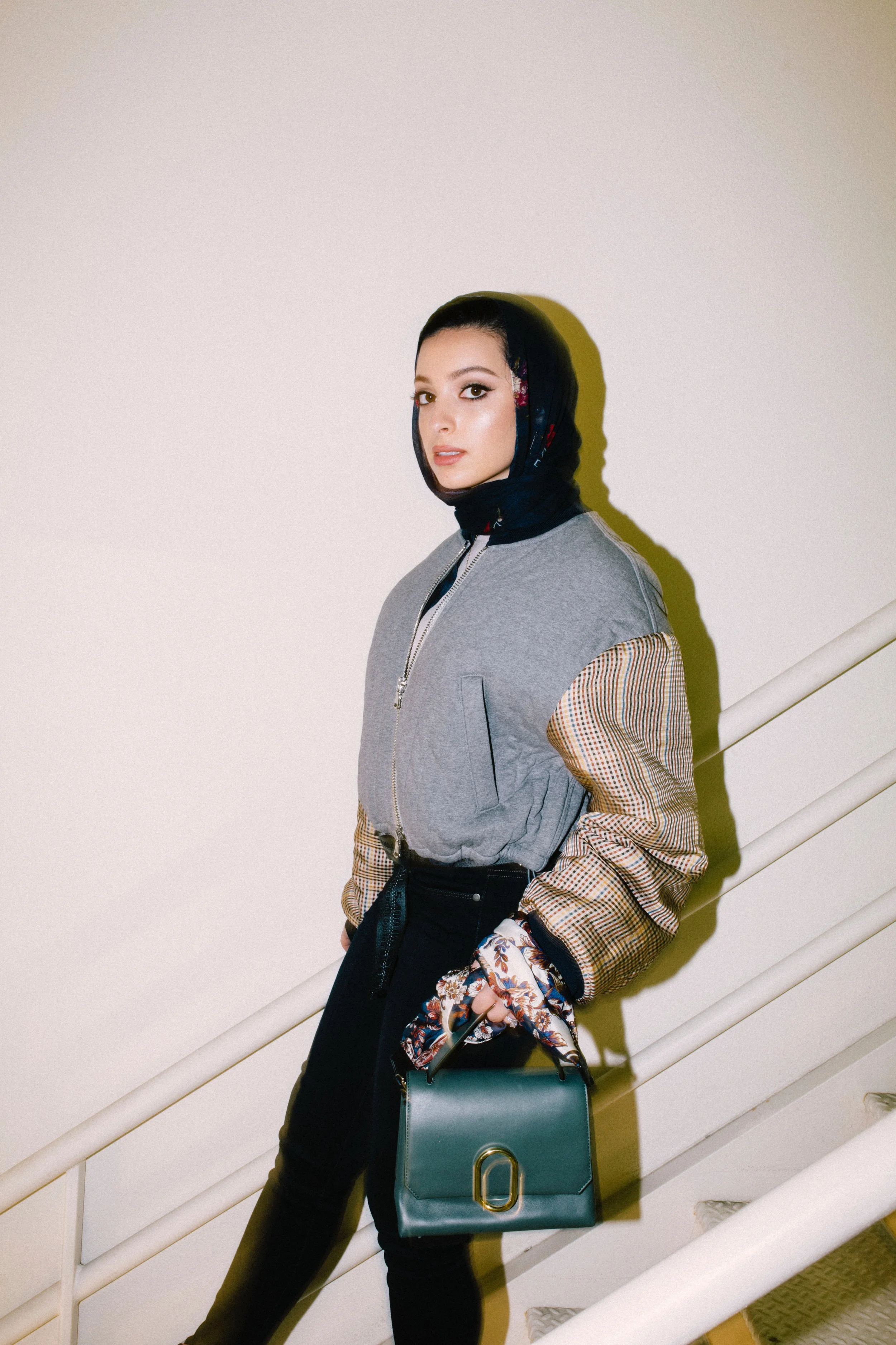 A woman standing on a staircase against a plain wall. She has dark hair, is wearing a black headscarf, a gray jacket with plaid sleeves, black pants, and carries a teal handbag.
