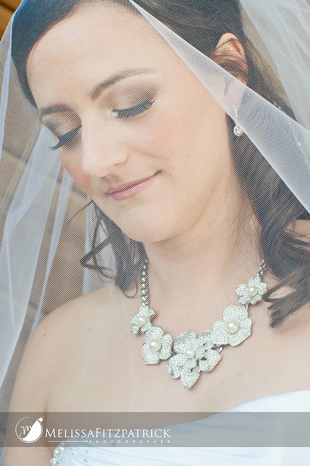 Close-up of a bride with a veil, wearing pearl and floral jewelry, with makeup and hairstyle, looking down with a smile.
