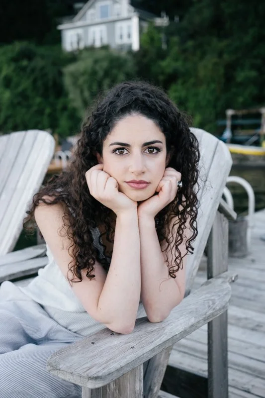 A young woman with curly dark hair leaning on her elbows on a wooden bench outdoors, resting her face in her hands, looking directly at the camera with a serious expression. Background includes a house on a hill, trees, and water with boats.
