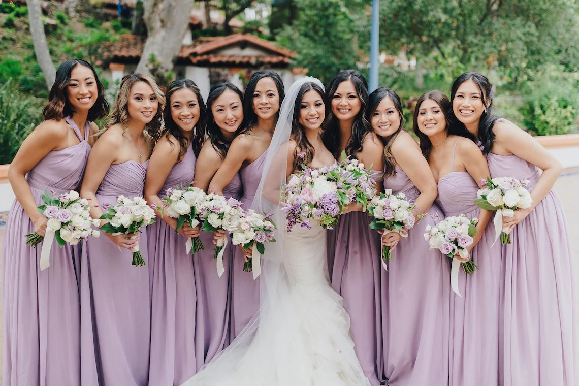 A bride and nine bridesmaids posing outdoors on a wedding day, holding bouquets of white and purple flowers, dressed in lilac dresses, with trees and a building in the background.
