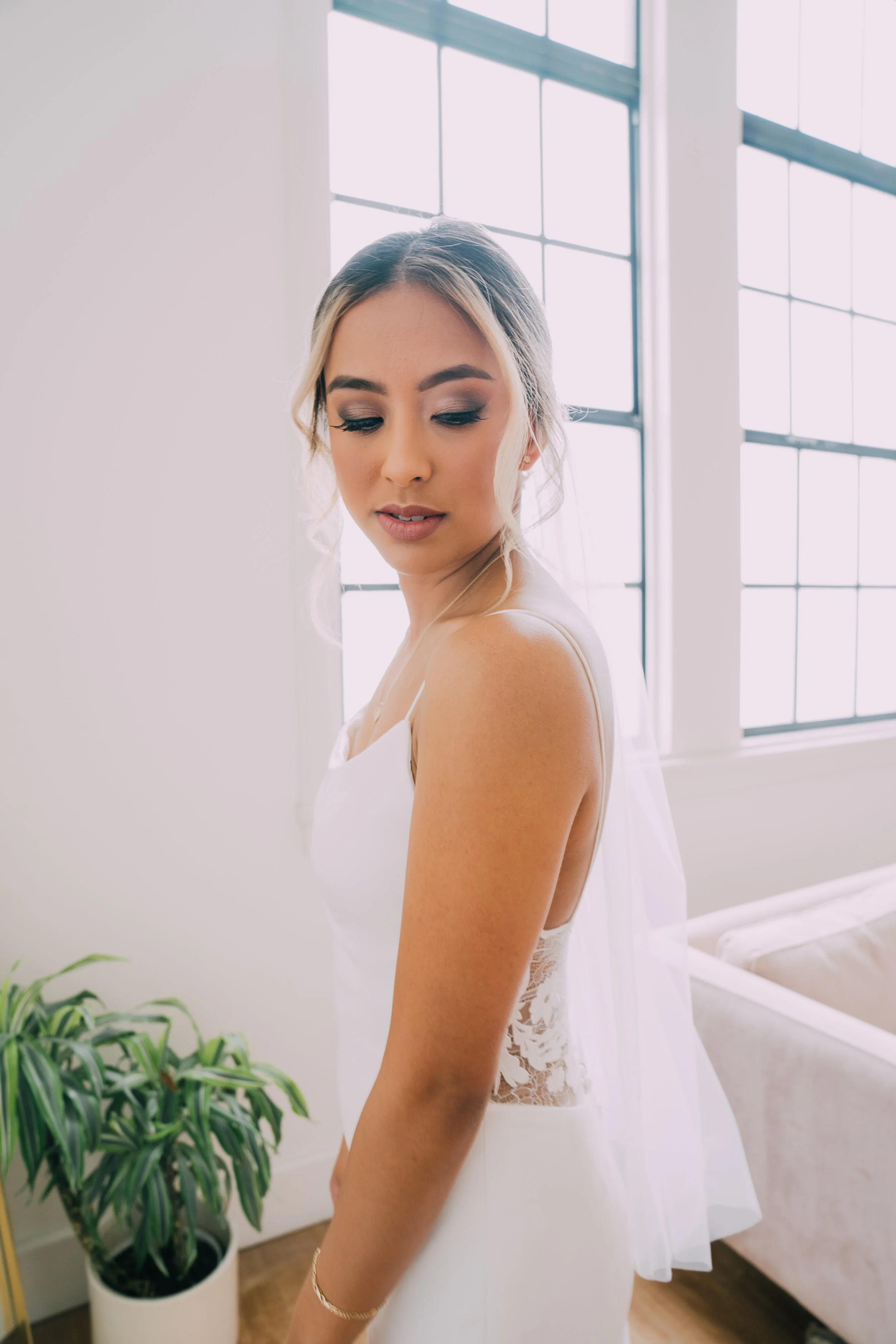 A young woman in a white dress with lace details looking down and to the side near a large window with grid panes, in a bright room with a potted plant in the foreground.