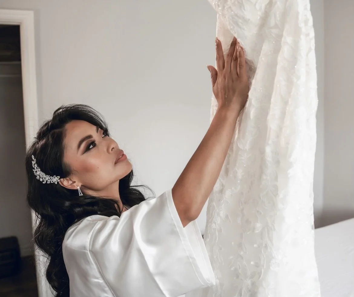 A woman with dark hair and jewelry, dressed in a white satin robe, looks at a white wedding dress hanging in front of her, with her hand touching the fabric.