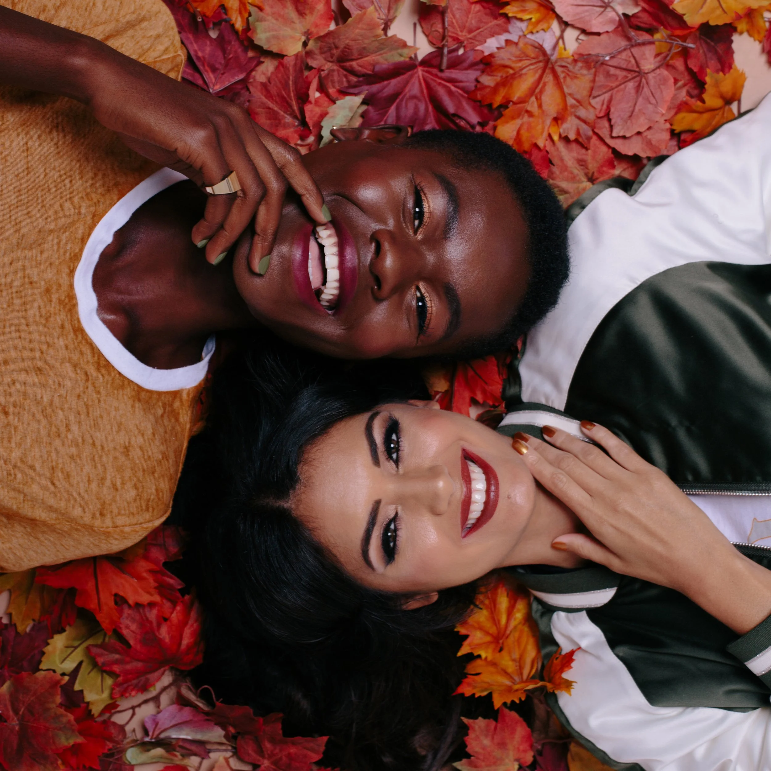 A woman with a joyful expression lying on a bed of colorful fall leaves, surrounded by her friends, all smiling and having fun.