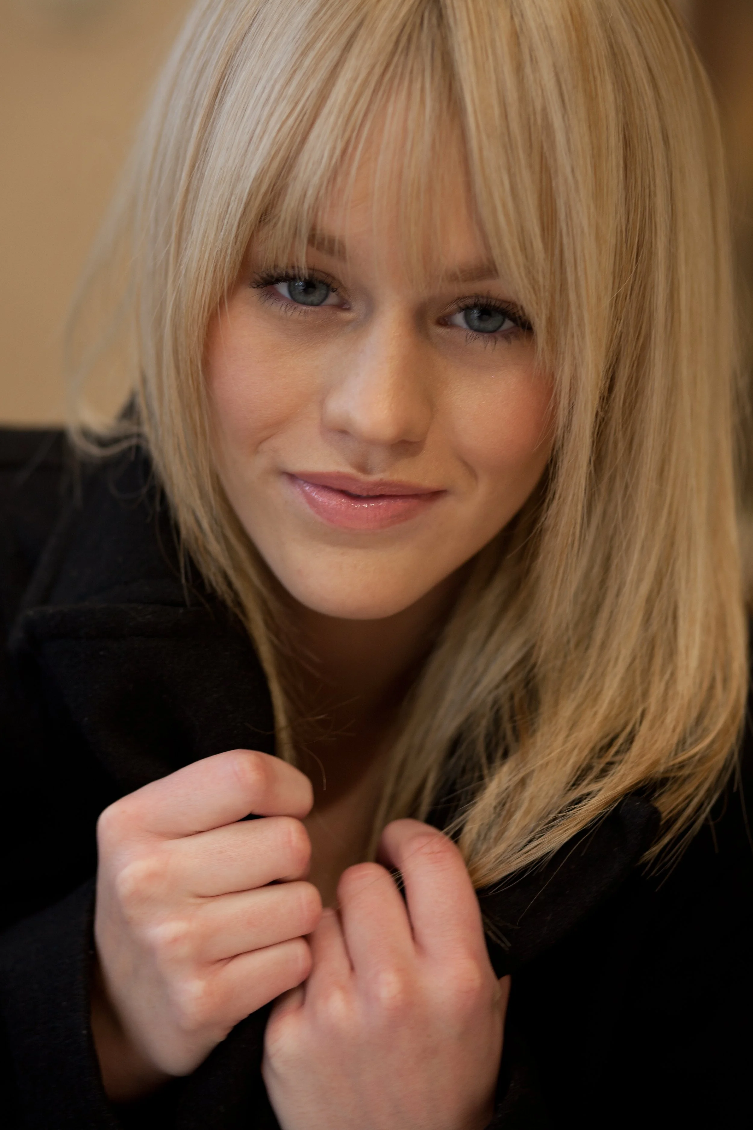 A woman with blonde hair and blue eyes, wearing a black top, looking at the camera and holding her hands near her chest.