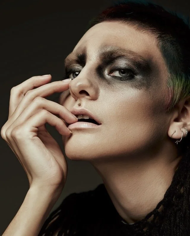 Close-up portrait of a person with bold makeup, short dark hair with colorful accents, and a nose piercing, posing with fingers near lips against a dark background.