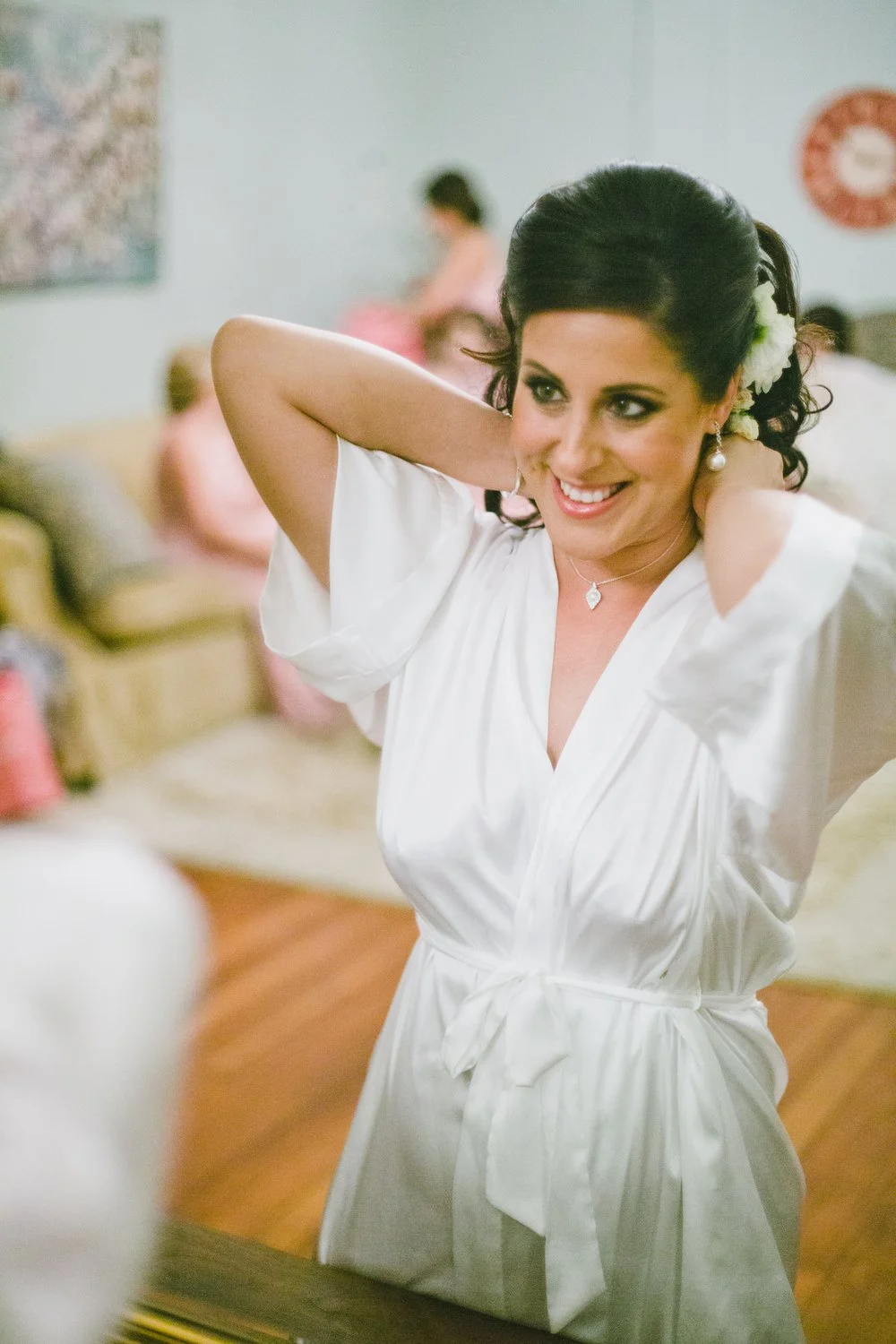 A woman in a white robe smiling as she adjusts her hair, with others in the background in a cozy room.