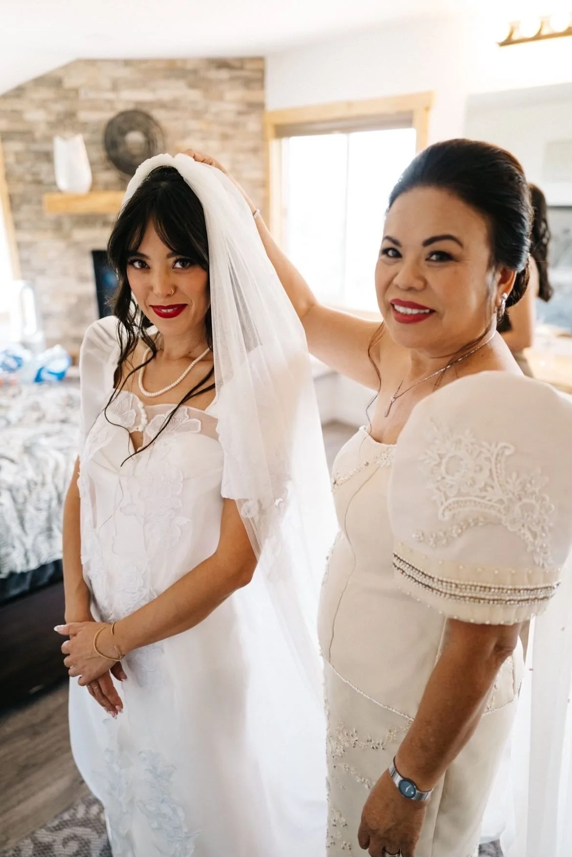 A woman helping another woman try on a wedding veil. The bride has dark hair, red lipstick, and is wearing a white dress and pearl necklace. The woman assisting is smiling, wearing a white dress with lace details, and has a watch on her wrist. They a