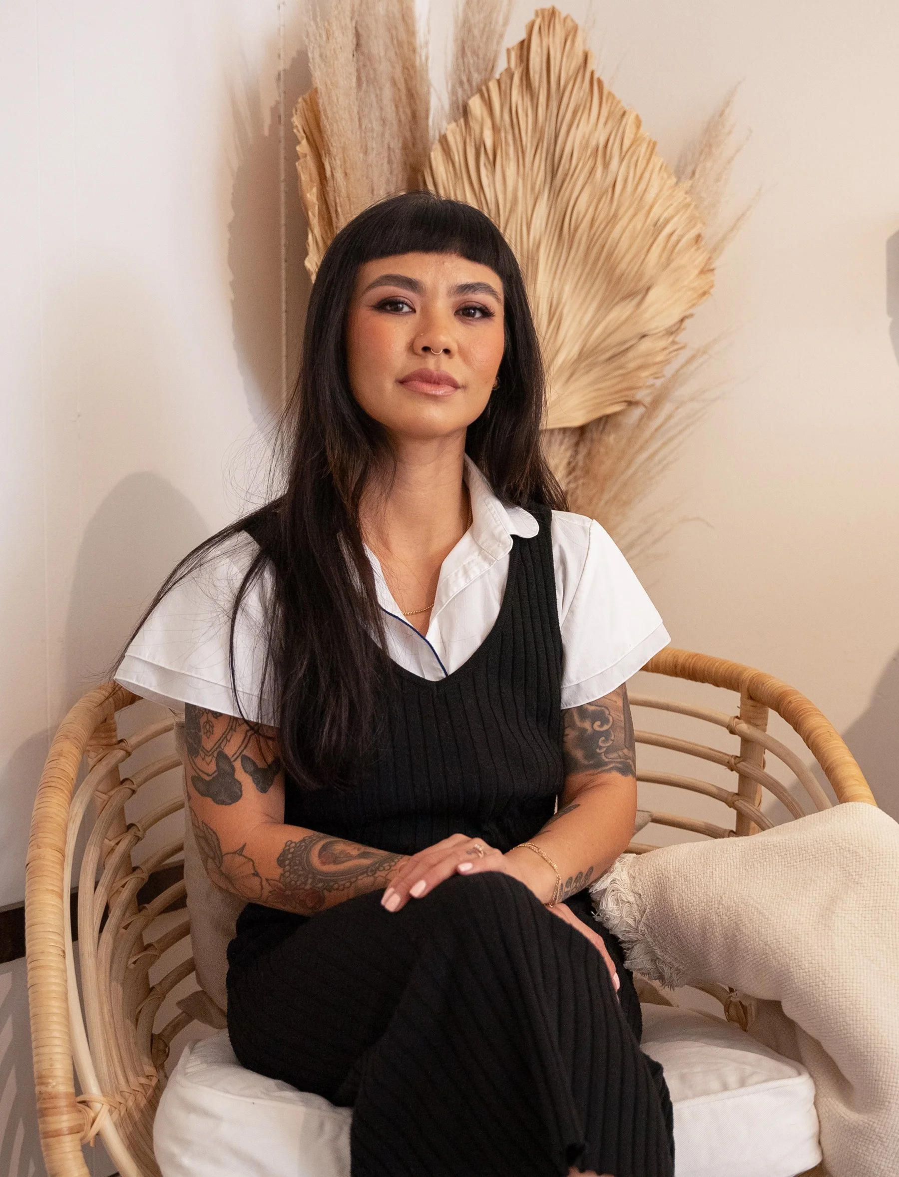 Rachelle sitting on a rattan chair with a cushion and beige blanket, against a white wall with large dried palm leaves or ferns in the background.
