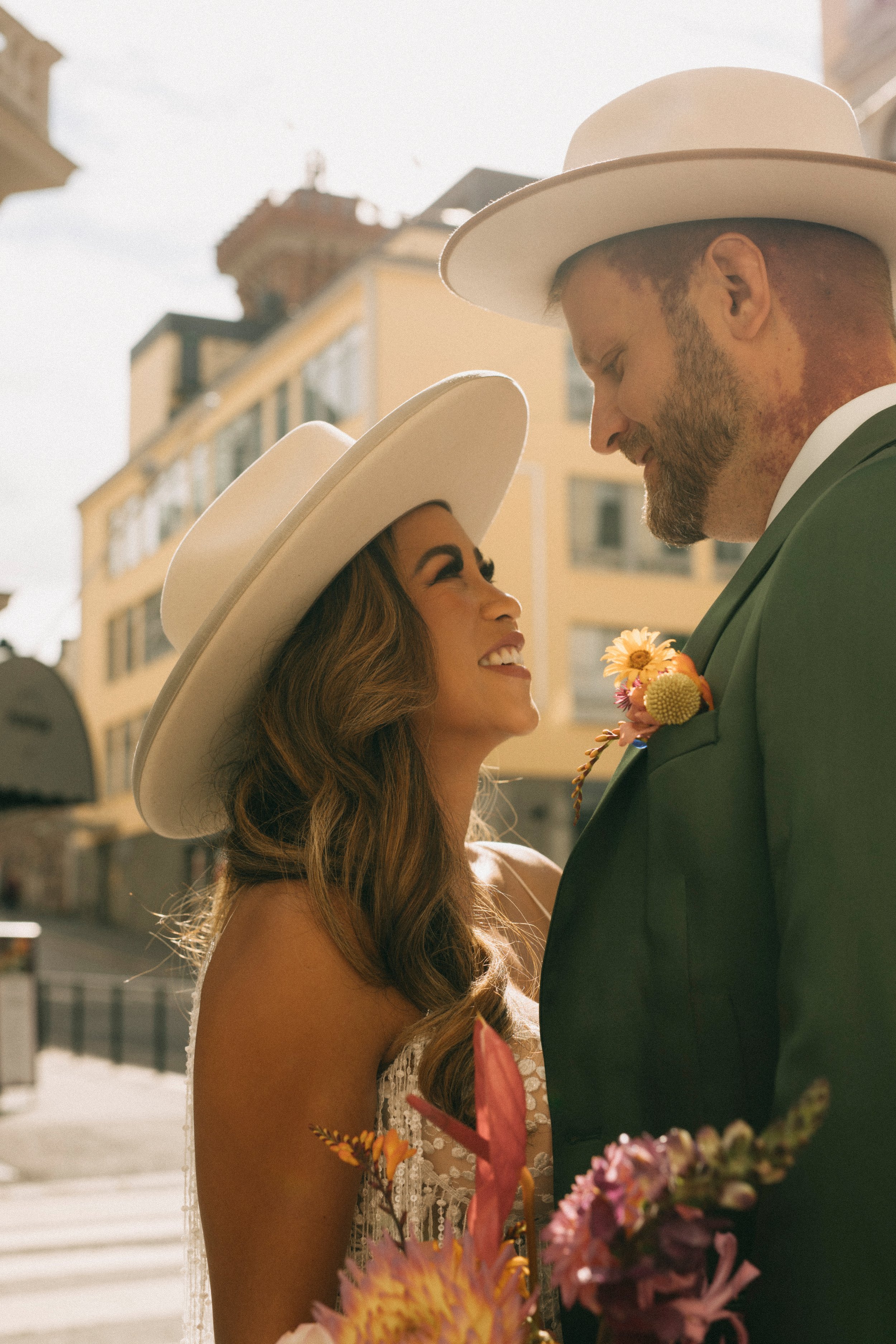 A man and woman in wedding attire wearing white wide-brimmed hats, smiling at each other, holding bouquets of flowers, in a city street with buildings in the background.