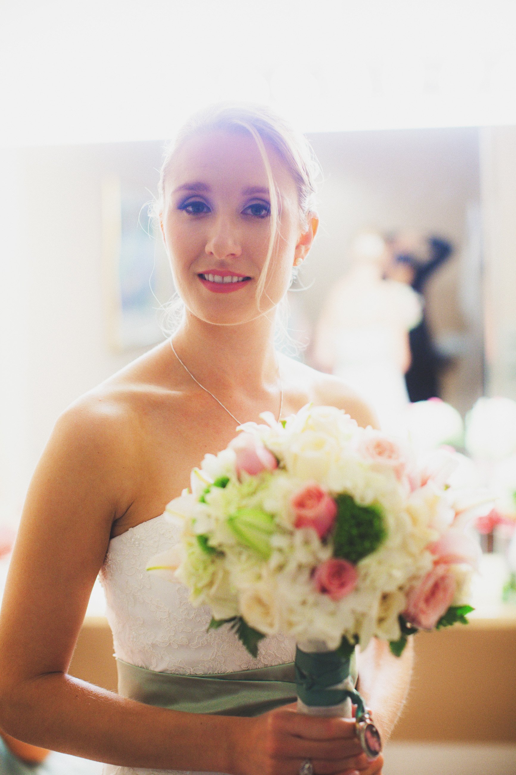 A bride holding a bouquet of pink and white roses, wearing a strapless white wedding dress, with her blonde hair styled up. The background is bright and slightly out of focus.